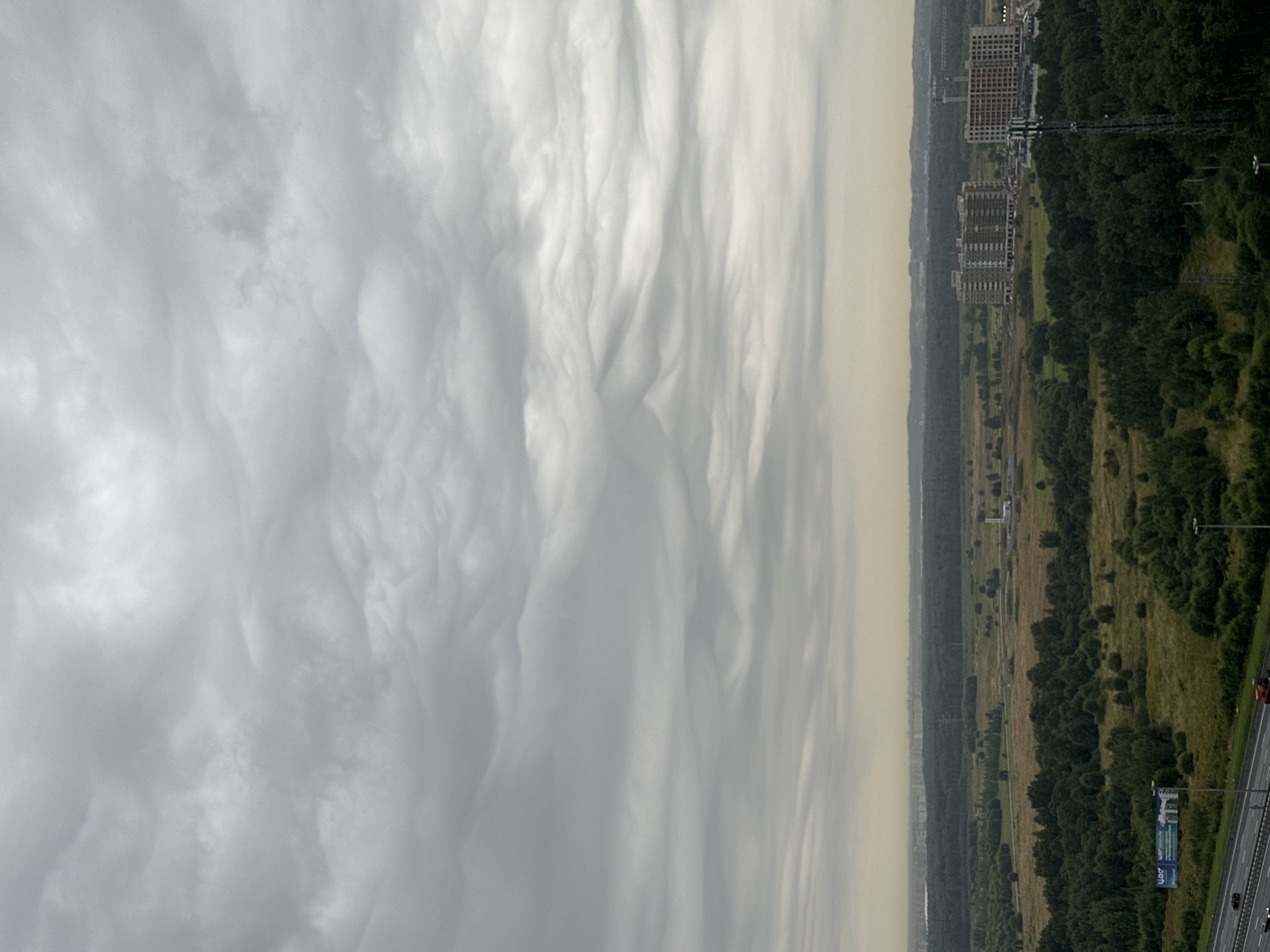 The image shows a wide, overcast sky with turbulent cloud formations. Below the sky, a landscape unfolds with a mix of forest, fields, and urban development. In the foreground, a multi-lane highway is visible with a few vehicles. A large billboard advertising a real estate development is present. Further into the landscape, clusters of trees and open grassy areas are seen. In the middle ground, there are several multi-story apartment buildings under construction and a power pylon. In the distance, a hazy horizon suggests a city or town. The location is identified as P.im.sverdlova, Russia. There are no discernible people or direct interactions captured in the image. The overall impression is one of an urban fringe environment under a dramatic sky.