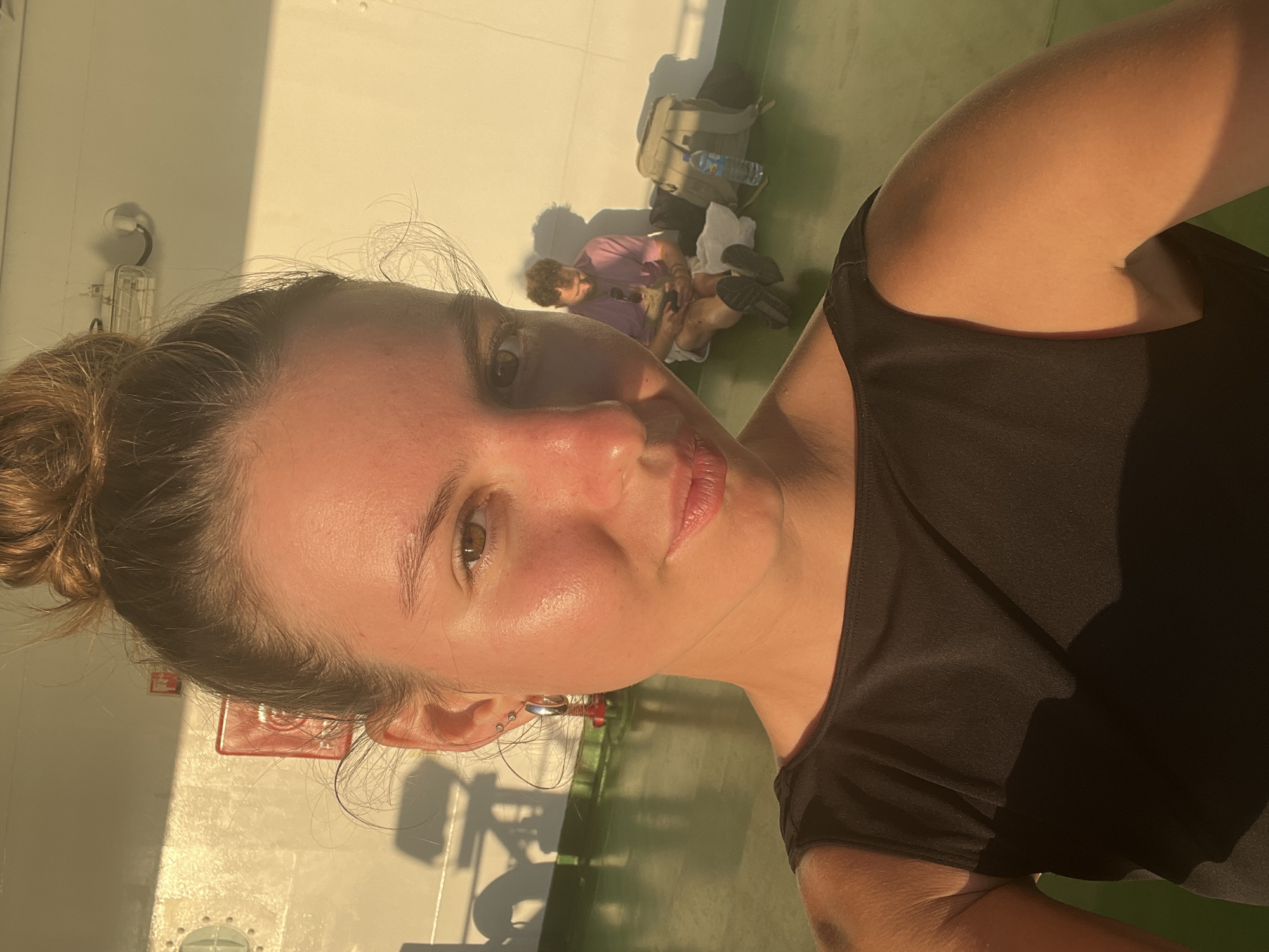 The image shows a young woman with her hair in a bun, looking directly at the camera. She is wearing a black sleeveless top and large hoop earrings. Her skin is illuminated by warm, golden light. In the background, out of focus, a man is sitting on the floor, looking at his phone. He is wearing a purple shirt and shorts. There is a backpack and a water bottle next to him. The environment appears to be an outdoor deck area with a white wall and a green floor. The scene is bathed in the warm glow of what appears to be sunset or sunrise. The image is a selfie taken by the woman in the foreground. The location is not specified as Batas Desa Skotong Timur, Indonesia.