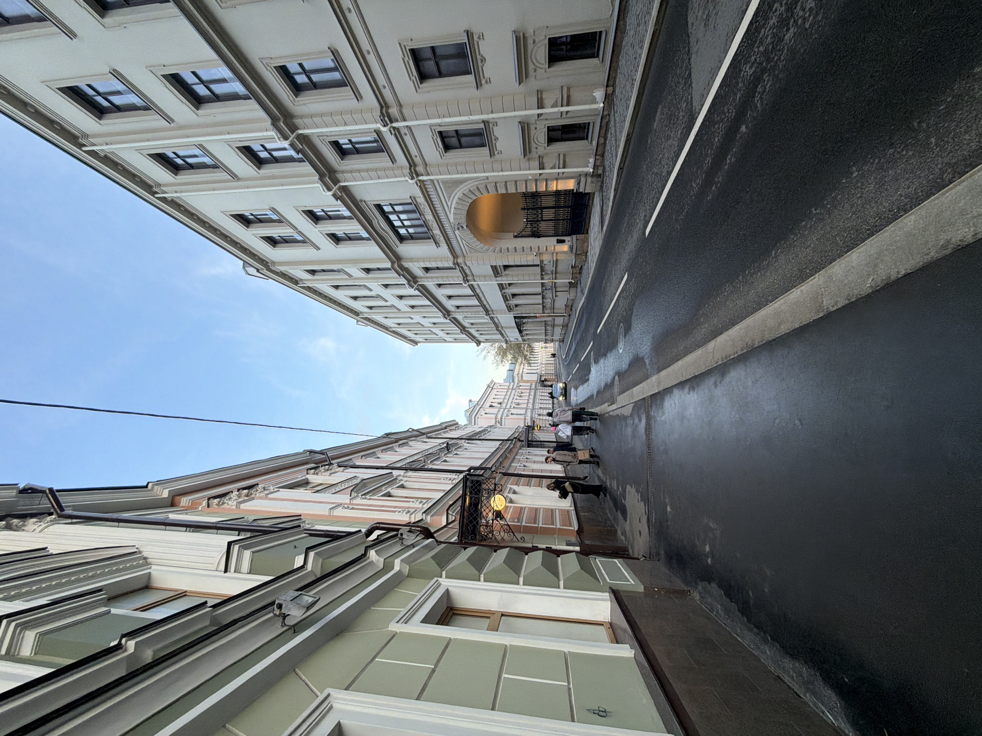 A street view, taken with a wide-angle lens, looks upward toward the sky between buildings in Moscow, Russia. The street appears wet and reflects the buildings above. A group of people walk along the sidewalk on the right side of the street. The buildings feature architectural details like windows, moldings, and cornices. One building has an arched gateway. A utility line stretches diagonally across the sky.