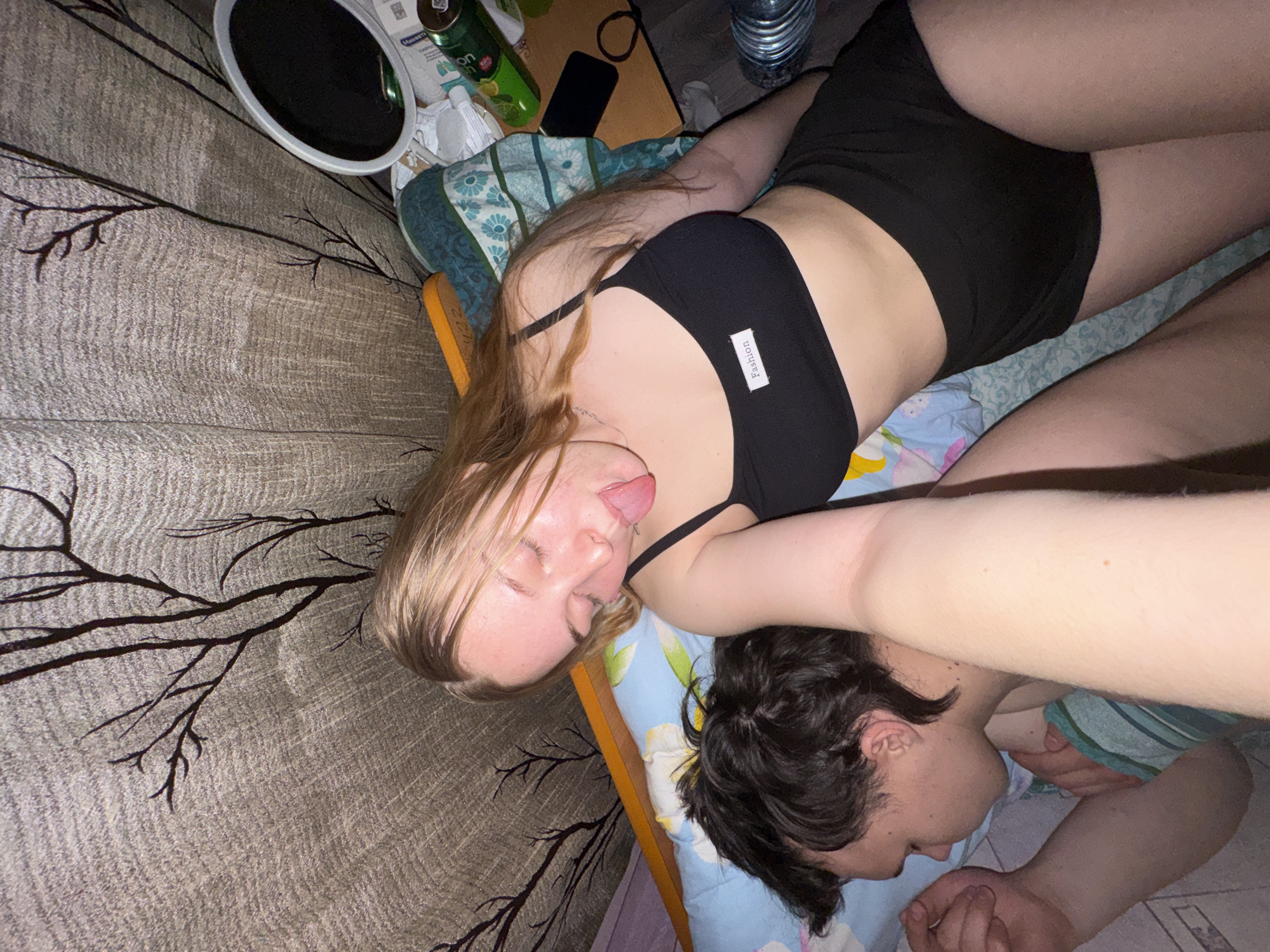 The image depicts two young adults in an indoor setting, likely a bedroom. The specific location in the city of Perm, Russia, cannot be determined from the visual information provided.
In the foreground and center, a female individual with long, light-colored hair is visible from the chest up. She is wearing a black sports bra with a white label reading "Fashion" and black shorts. Her eyes are closed or squinted, and her tongue is extended playfully. Her right arm is angled towards the viewer, suggesting she is holding the camera.
To her left, partially obscured by her arm and head, a male individual with dark, disheveled hair is lying on his side, facing away from the camera. He appears to be sleeping or resting, covered by a light blue sheet with a floral or patterned design.
The bed is covered with patterned bedding, including a light blue sheet with cartoon-like elements and a teal-green blanket with white floral patterns. An orange hair care item, possibly a comb handle, is visible near the female's head.
The background on the right side of the image reveals a cluttered bedside surface. Objects present include a small, round, silver-rimmed mirror, at least two green beverage cans (one identifiable as Lipton Iced Tea), various small bottles and containers (one appears to be Miramistin), a black smartphone, and a black hair tie. On the floor next to the bed, a ribbed plastic water bottle is visible.
The wall or curtain behind the subjects features a beige or light brown pattern resembling tree branches. The overall lighting is dim, suggesting an evening or low-light environment.