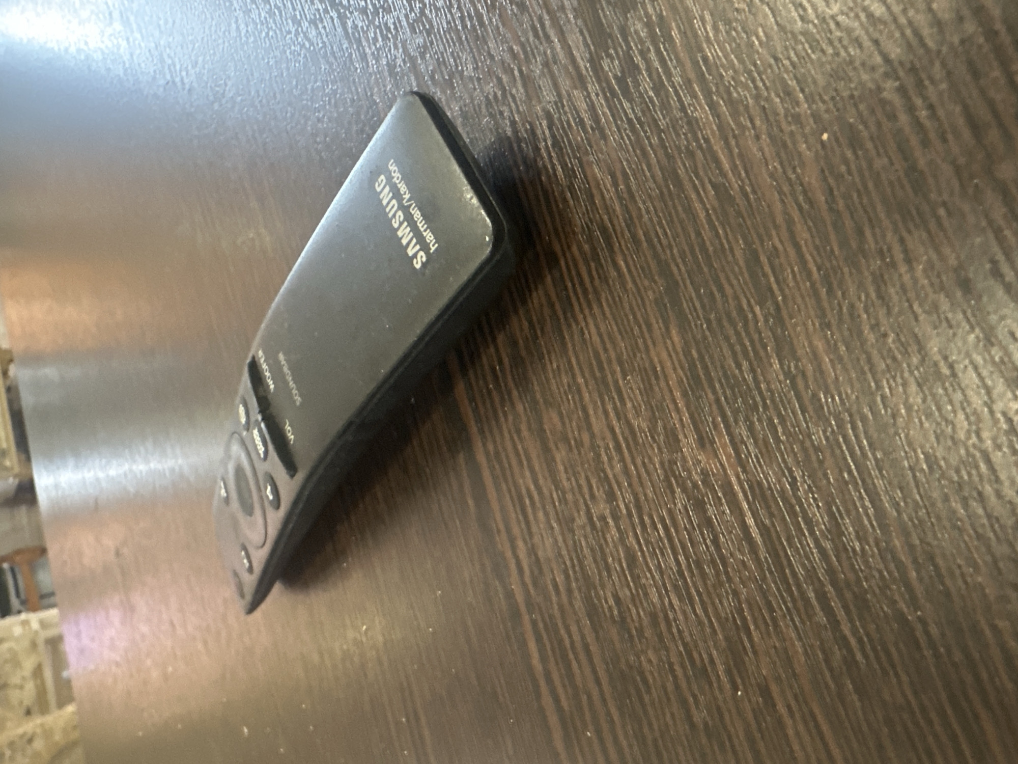 A black Samsung soundbar remote control with "harman/kardon" branding lies on a dark brown wooden table. The table has visible wood grain and some light-colored specks, possibly dust. The remote control is angled with its top-left corner closer to the camera. It has several buttons, including a circular directional pad and smaller buttons labeled "VOL" and "WOOFER." The lighting in the scene is bright and appears to be coming from the left, casting a slight reflection on the table's surface. The background is out of focus, showing what appears to be furniture. The scene is located in the city of Artemovsky, Russia.