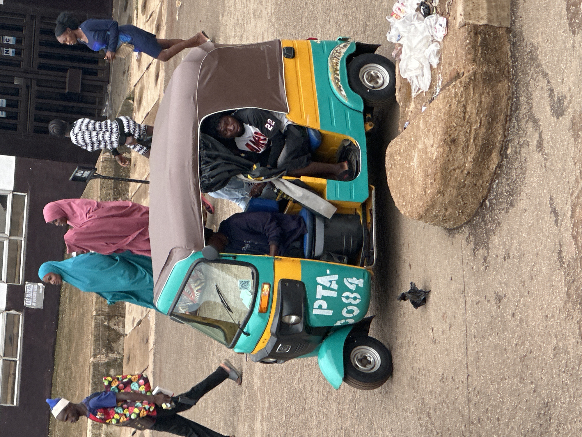 In Bukuru, Nigeria, a bright teal and yellow auto-rickshaw is parked on a paved road. The rickshaw, identified with the markings "PTA-3084," has a brown canopy. Inside, a young man wearing a black and white jersey with the number "22" on it is seated, looking towards the left. Another person is partially visible in the driver's seat.
Several people are walking in the background. To the left, a person in a blue shirt and a white and blue hat walks with a colorful bag. Further back, two women in brightly colored hijabs, one teal and one pink, are walking. To the right of them, a woman in a black and white patterned dress walks, followed by a woman in a dark blue dress. A black bird is on the ground in front of the auto-rickshaw. The environment includes a rough, rocky structure to the right of the vehicle, with some scattered plastic bags and debris on it. The background shows a building with barred windows and signage.