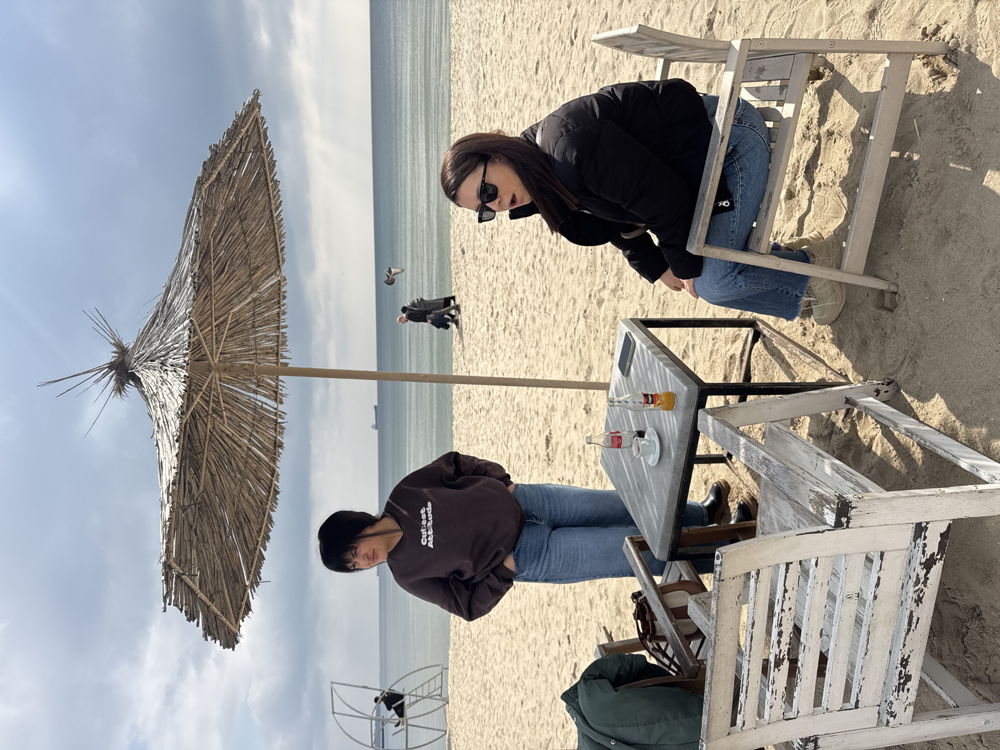 Two women are seated at a white, weathered table on a sandy beach. One woman, wearing a brown sweatshirt with the words "Cutest Attitude" and blue jeans, has short dark hair and is leaning on the table with her hands in her pockets. The other woman, wearing a black jacket, blue jeans, and sunglasses, is leaning forward in her chair. On the table are bottles, a cup, and a small plate. An umbrella made of straw provides shade for the table. In the background, the sea stretches out to the horizon under a cloudy sky. Some people are walking along the beach in the distance. A ladder-like structure is visible in the lower left corner. The setting is likely near Varna, Bulgaria.