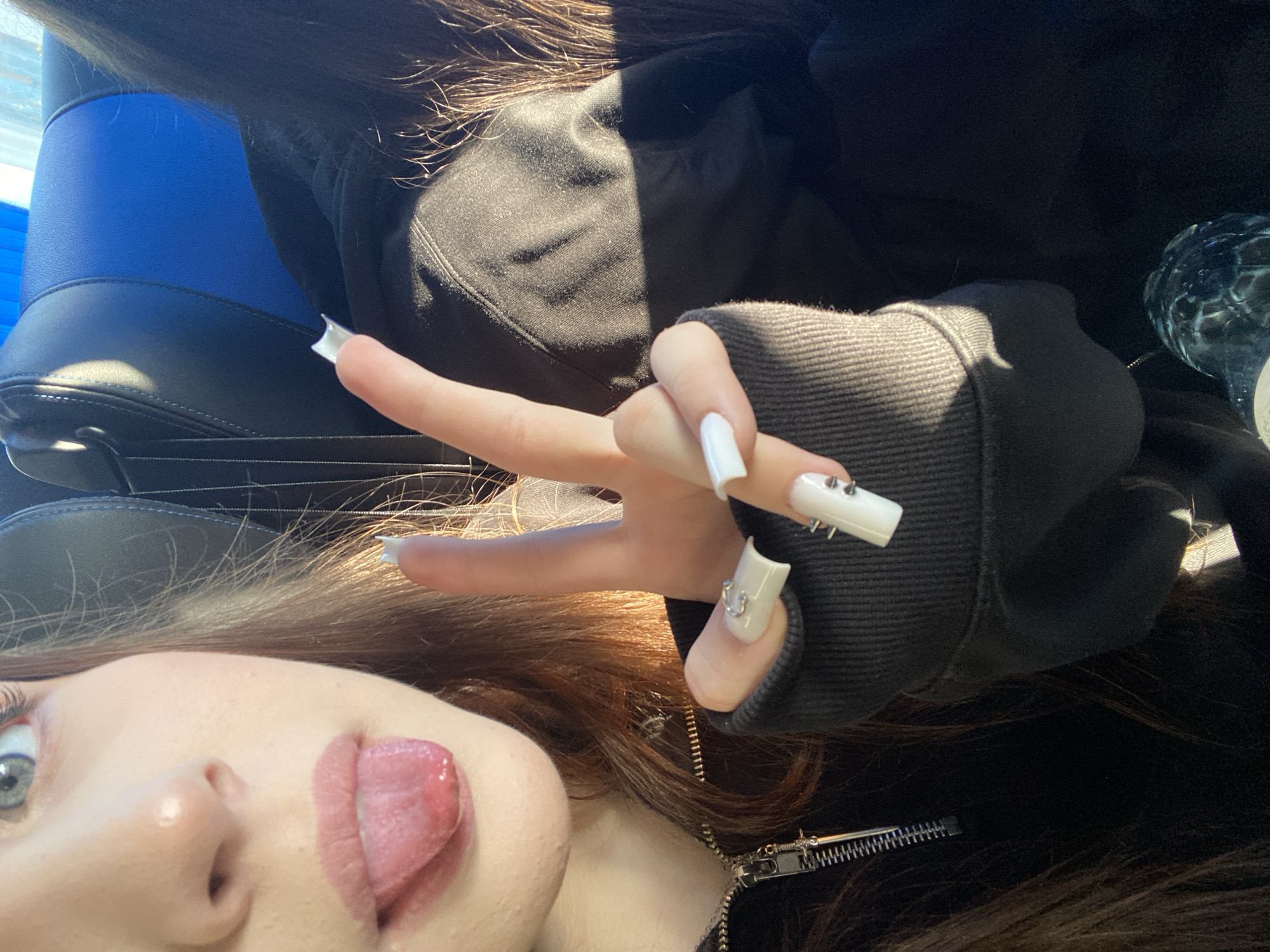 A close-up selfie shows a young person sticking their tongue out. The subject has long brown hair and fair skin. Their right hand is raised, forming a peace sign, with long white fingernails adorned with studs and a heart charm. They wear a black hooded sweatshirt with a visible zipper. The background includes a blue and black car seat. The location is not discernible from the image, thus the city of Moscow, Russia cannot be confirmed.