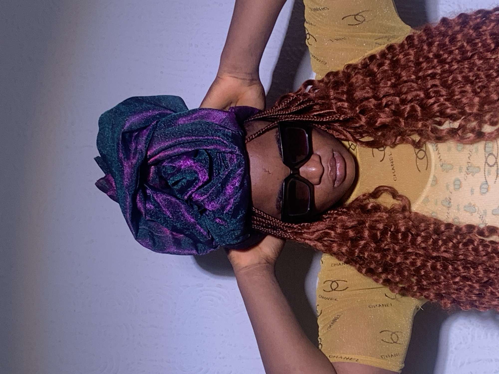 This close-up image features a stylish person, likely female, with dark skin, lying down and looking upwards towards the camera. Her head is positioned towards the left side of the frame. She wears large, rectangular dark sunglasses that obscure her eyes, and her lips are slightly parted. A striking headwrap, made from a vibrant purple and dark green metallic fabric, is elegantly tied on her head, forming a voluminous, rose-like shape on the left side.
Her hair consists of long, reddish-brown, tightly coiled curls, which cascade around her head and upper body. Some braided sections are visible near her face. She is dressed in a sheer, shimmering yellow long-sleeved top that is patterned with numerous black "CHANEL" text and interlocking double 'C' logos. Her arms are raised, with her hands appearing to gently cup or frame her head and headwrap. The background is a plain, light grey or off-white surface, indicating an indoor setting. The overall composition suggests a confident and fashionable pose. The scene is set in Barakin Akawo, Nigeria.