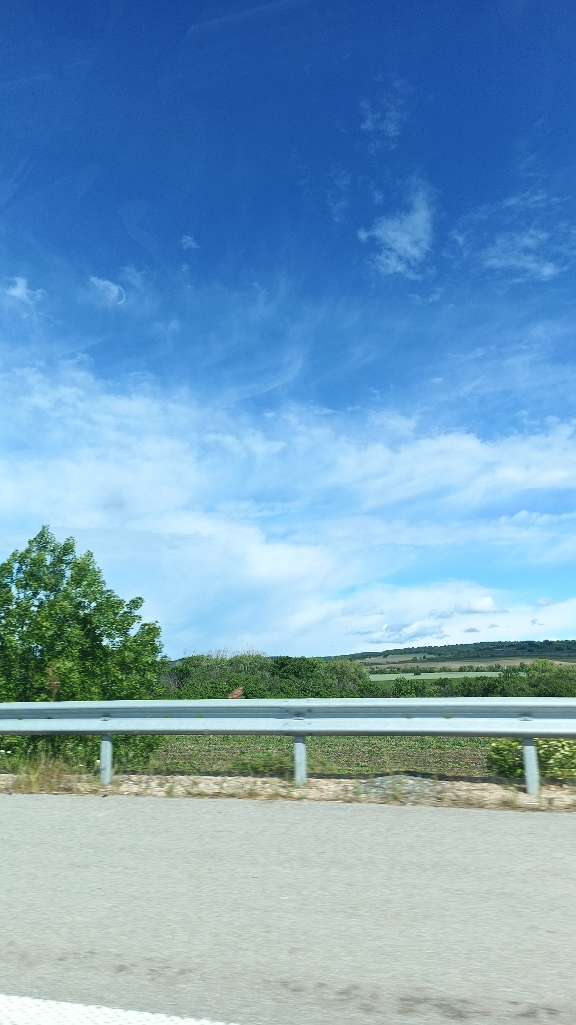 A daytime, outdoor scene shows a road with a gray guardrail running along the edge. Beyond the guardrail, there is a green field with low-lying vegetation and trees. Further in the distance, there is a hilly landscape with patches of trees and fields. The sky is blue with scattered white clouds. No people or other discernible subjects are present. The location details are Yazdach, Bulgaria.