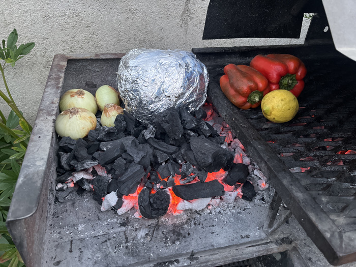 A charcoal grill contains vegetables being cooked. There are four onions, two red peppers, and one lemon atop the grill. A food item is wrapped in aluminum foil and nestled among the hot coals. Black charcoal pieces surround the vegetables. The grill is dirty and gray.