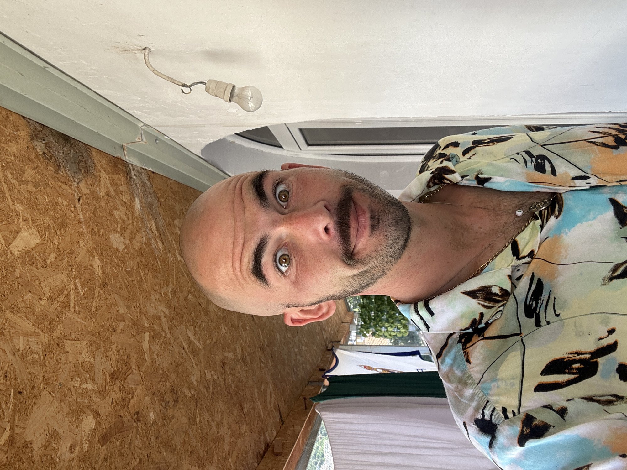 A bald, bearded man with green eyes is looking at the camera. He is wearing a patterned shirt with gold chain. The background consists of a ceiling with an exposed light bulb and a window. Part of the plywood wall is visible on the left. Outside the window, a glimpse of greenery and buildings can be seen. While the specific location within Budva, Montenegro cannot be determined from the image, the overall environment suggests an interior space with access to an outdoor area within an urban setting.