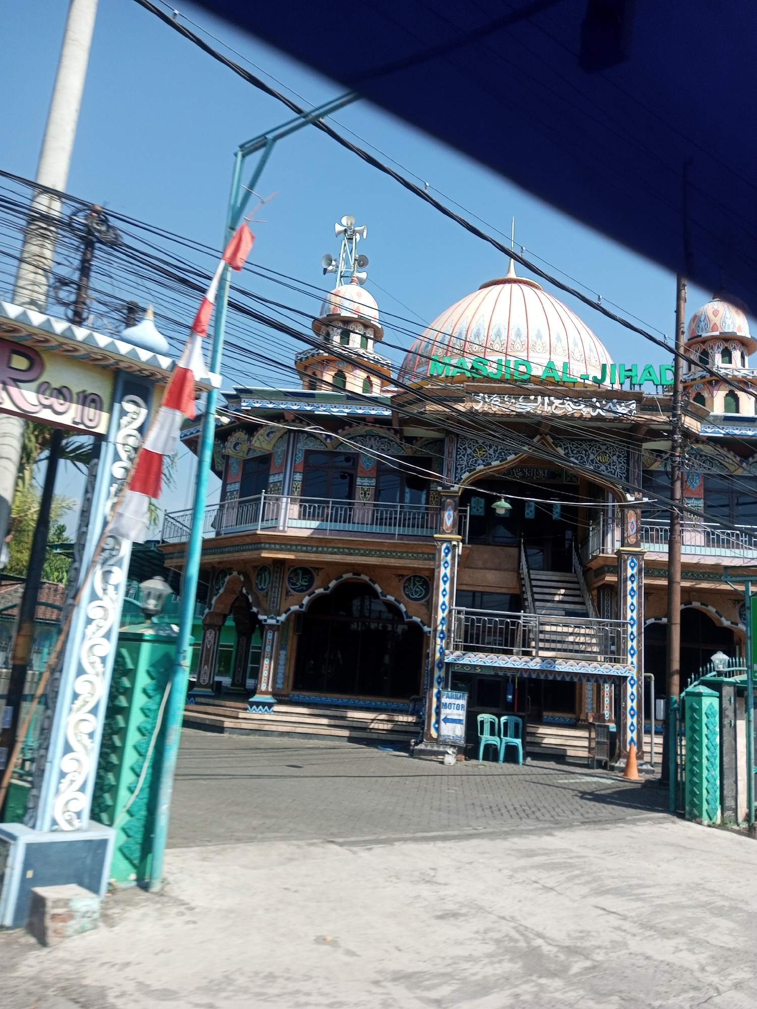 The image shows the exterior of Masjid Al-Jihad in Karangsono, Indonesia, on a clear day. The mosque features a large dome adorned with intricate patterns and Arabic calligraphy, along with smaller minarets. The facade is colorful with orange, blue, and white accents. In front of the mosque, there is an entrance with stairs and a balcony. To the left, a gate with decorative pillars and a red and white flag, likely the Indonesian flag, is visible. The area is cluttered with numerous electrical wires overhead. A sign with an arrow pointing left and the text "PARKIR MOTOR" (Motorcycle Parking) is present near the entrance. Two light blue plastic chairs are placed on the paved ground in front of the mosque. An orange traffic cone is also visible on the pavement.
