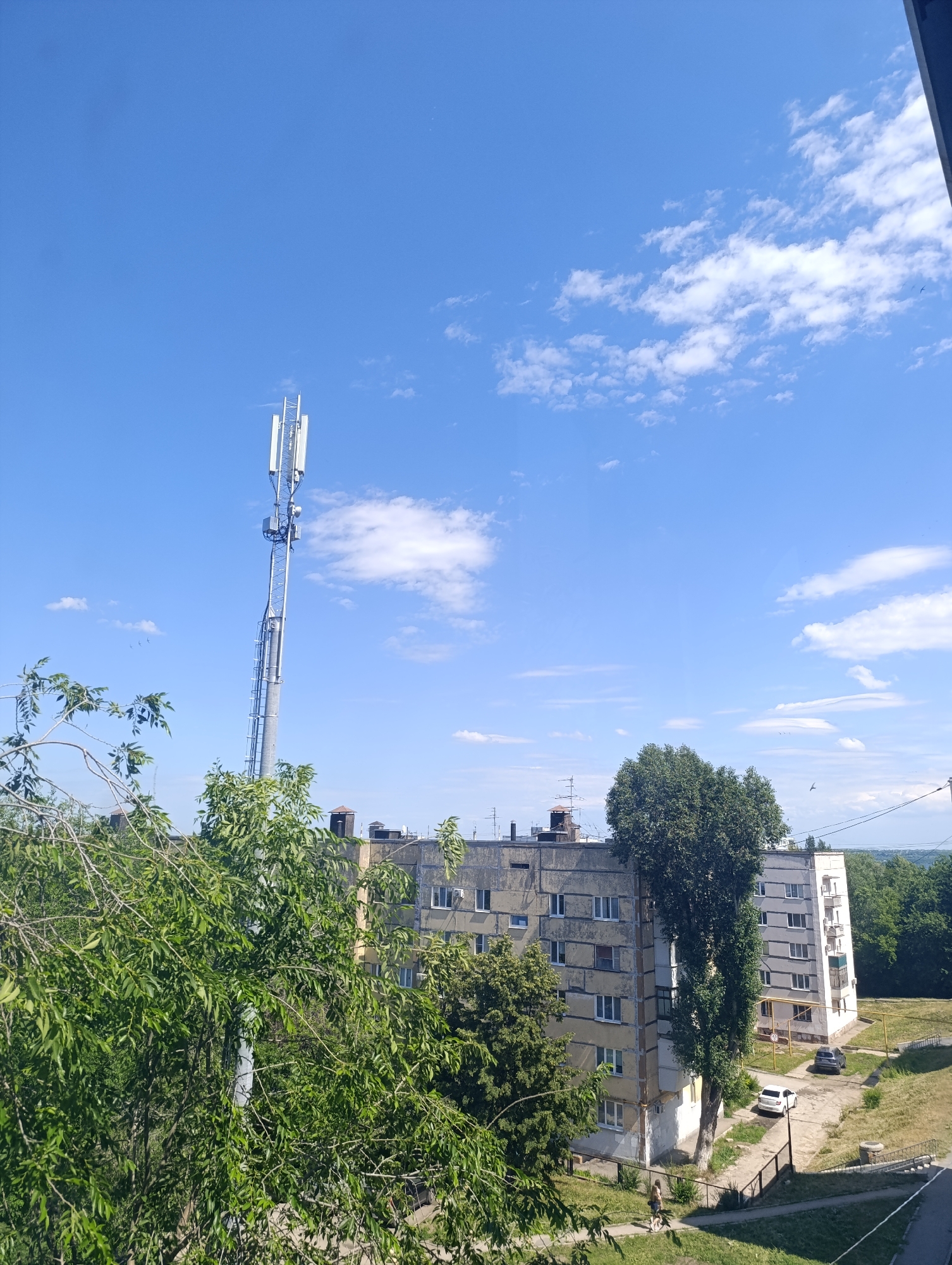 In an outdoor urban environment under a clear blue sky with scattered white clouds, a tall cell tower stands prominently in the left half of the frame. The tower is metallic and slender, topped with multiple antenna arrays. In the middle ground, a multi-story, weathered apartment building with a gray facade and white window frames stretches horizontally. Adjacent to it, a second, lighter-colored apartment building stands slightly to the right, also featuring numerous windows. Dense green trees partially obscure the buildings from view. A paved area in front of the apartment buildings shows two parked cars: a white sedan and a darker SUV. A pathway with a grassy verge and a small slope leads down from the buildings. Further in the background, more trees and the suggestion of distant hills can be seen.