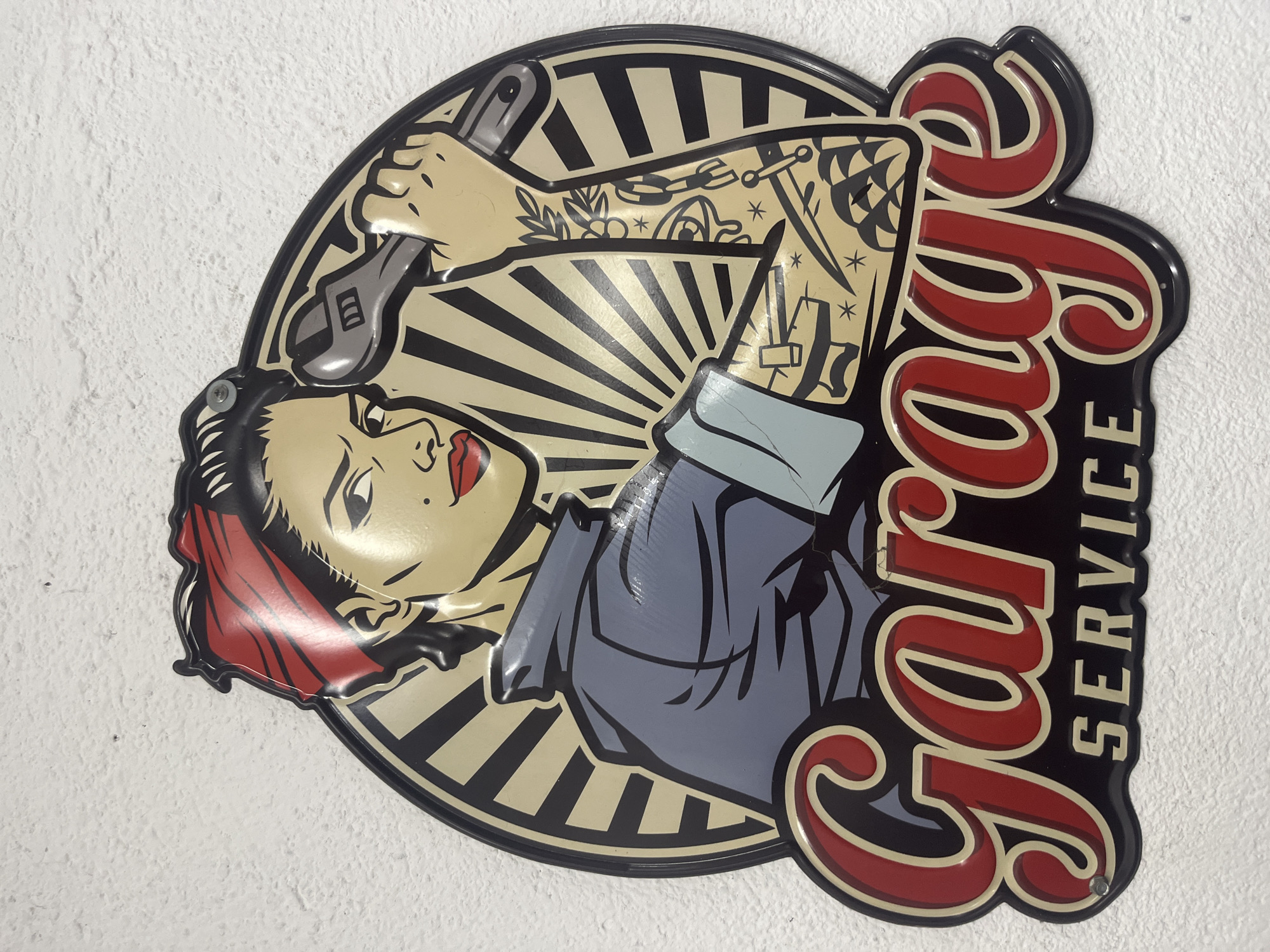 A metal sign depicting a stylized woman holding a wrench hangs on a textured white wall. The woman has dark hair tied with a red bandana and is wearing a blue shirt and light blue sleeves. Her left arm is covered in tattoos. The sign has the word "Garage" in large red and yellow lettering at the bottom, with "Service" written in smaller yellow letters below. The background of the circular part of the sign features radiating black and tan lines. The sign is mounted to the wall with visible screws. The image does not provide information about the city of Burgas, Bulgaria.