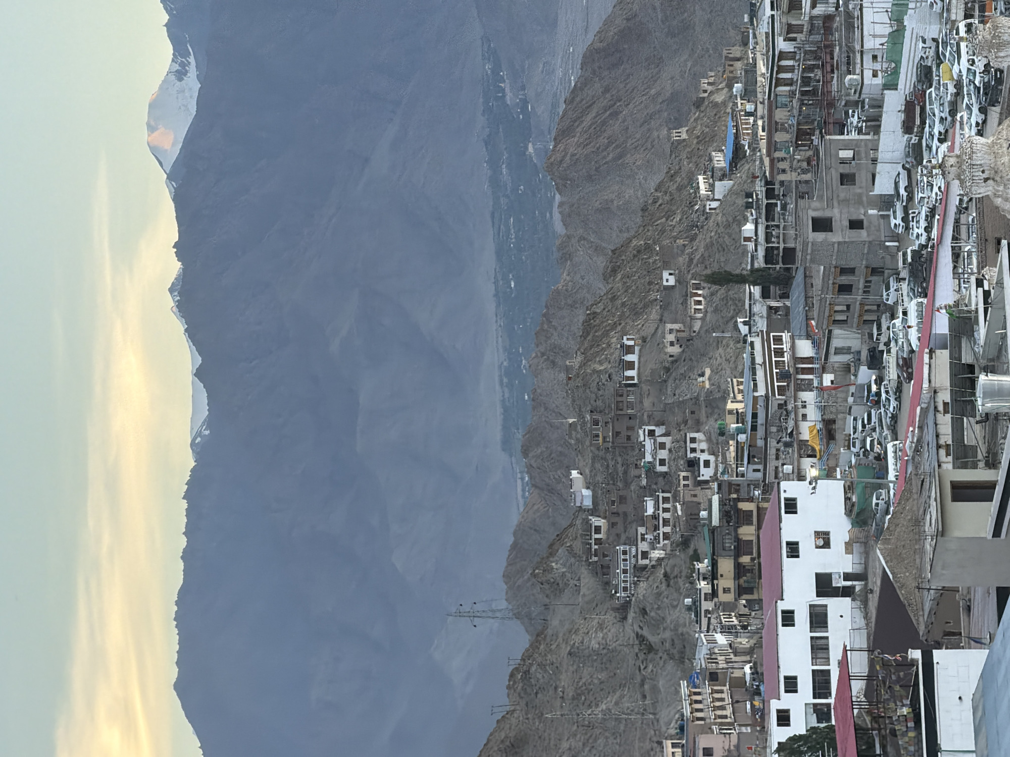 The image depicts a high-altitude town nestled in a valley with towering, rugged mountains in the background, characteristic of Leh, India. The sky above is a gradient of pale yellow and blue, suggesting either sunrise or sunset, with the peaks of distant snow-capped mountains catching the light. In the middle ground, numerous white-roofed, multi-story buildings are densely clustered on the steep, rocky slopes, their architecture reflecting the arid, mountainous environment. The houses are primarily constructed with stone and concrete, many featuring white walls and dark roofs. Some larger buildings, possibly hotels or administrative structures, are visible in the foreground, along with a significant number of parked vehicles, including cars and vans, indicating a populated area. Power lines and transmission towers are also present, a common sight in mountainous regions. The overall impression is of a remote, yet active, settlement set against a dramatic and imposing natural landscape.