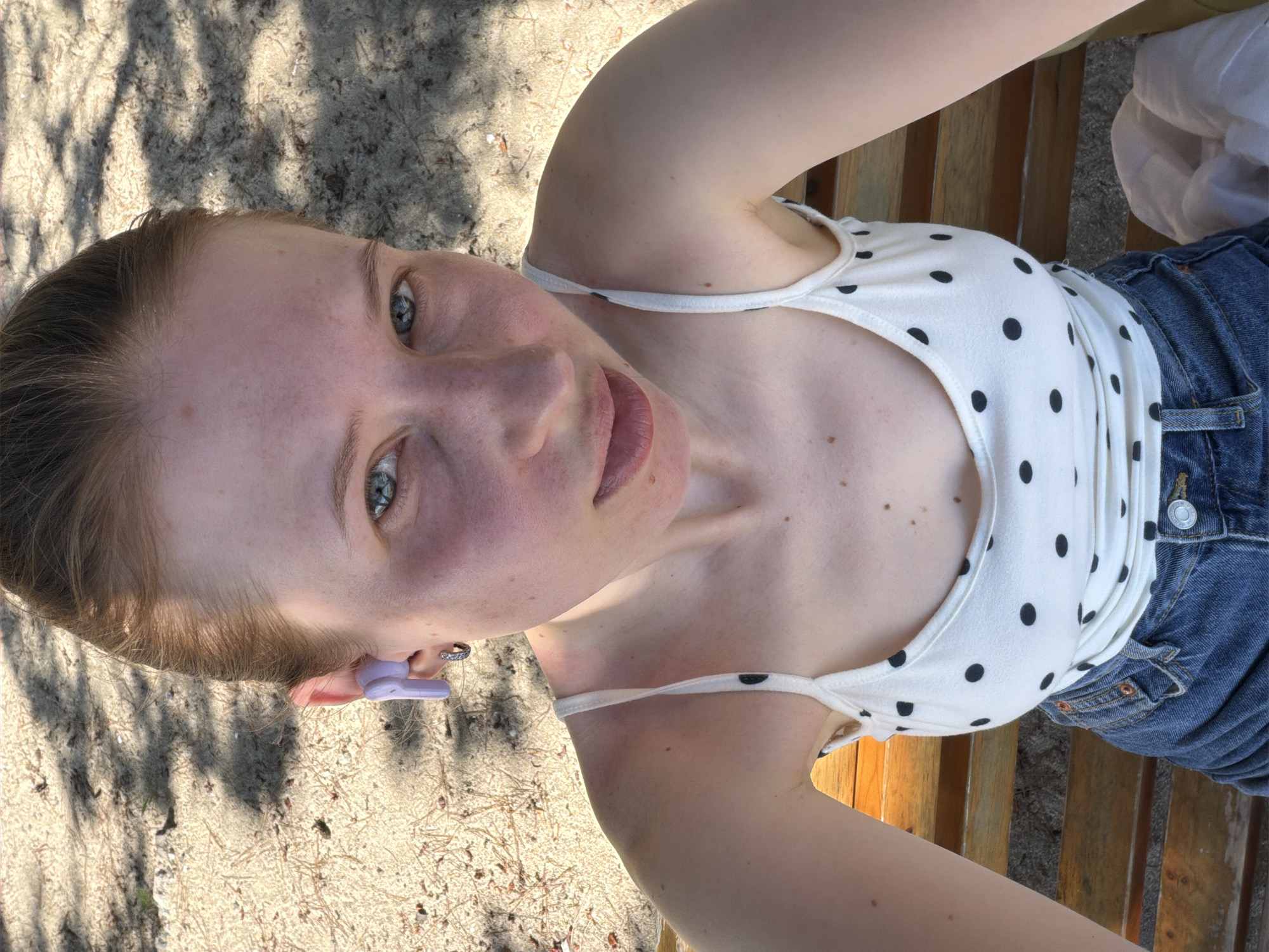A young woman with fair skin and freckles sits on a wooden bench outdoors. She has light brown hair pulled back and is wearing a white polka dot tank top and blue denim shorts. A single purple earbud is visible in her right ear, and she has a silver hoop earring in her left earlobe. The background consists of sand, with dappled shadows suggesting trees overhead. This scene is consistent with a public beach or park setting in Gangneung-si, South Korea.