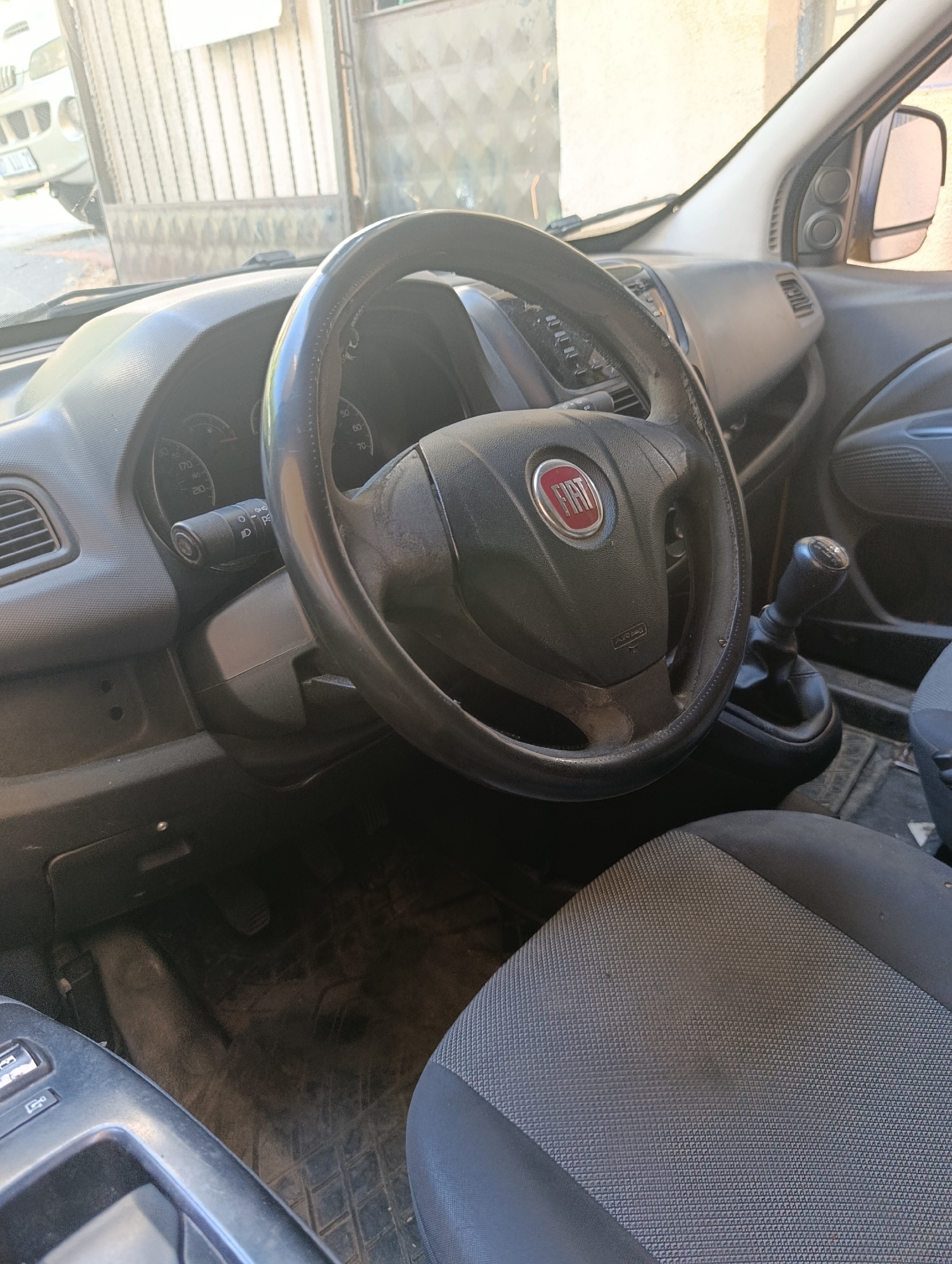 The image captures the interior of a car parked in Ümraniye, Turkey. The focus is on the driver's side, showcasing the steering wheel with the Fiat logo prominently displayed. To the right of the steering wheel is the gear stick, and the dashboard is visible, including the speedometer and various indicator lights. The car's interior appears somewhat worn, with the steering wheel showing signs of use. The passenger seat, upholstered in a grey fabric with a textured pattern, is also visible in the foreground. Through the windshield, an external environment is dimly discernible, suggesting the car is outdoors, possibly in a street or parking area in Ümraniye, Turkey. The lighting indicates it is daytime, with sunlight casting shadows and highlighting the car's interior elements. No individuals are present within the visible frame of the car.