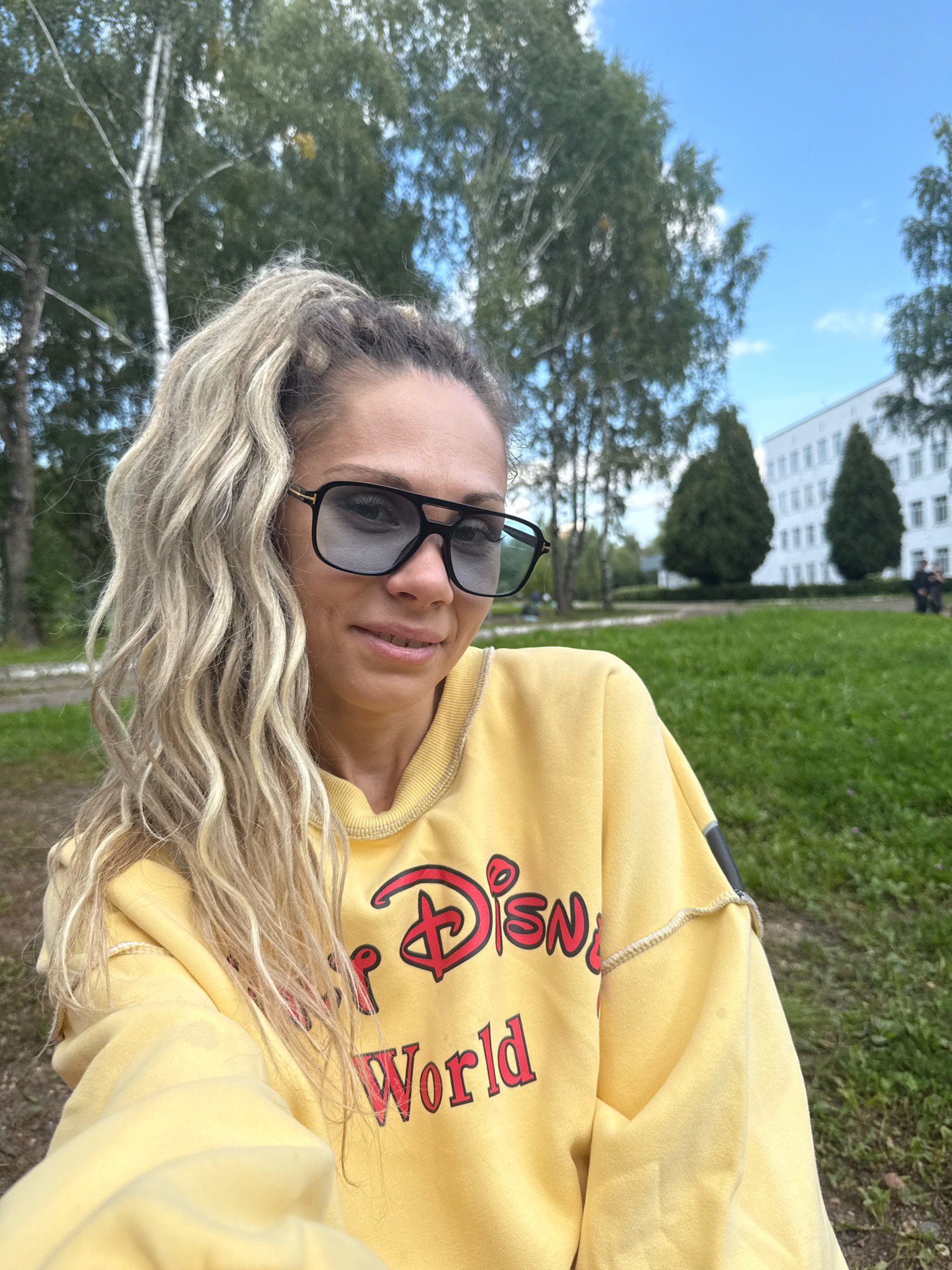 A woman with blonde, curly hair pulled into a high ponytail and wearing large, dark sunglasses is taking a selfie outdoors. She is smiling and wearing a yellow sweatshirt with a red Disney World logo on it. The background shows a grassy area with trees and a white building in the distance. The scene appears to be in the city of Rusanovo, Russia.