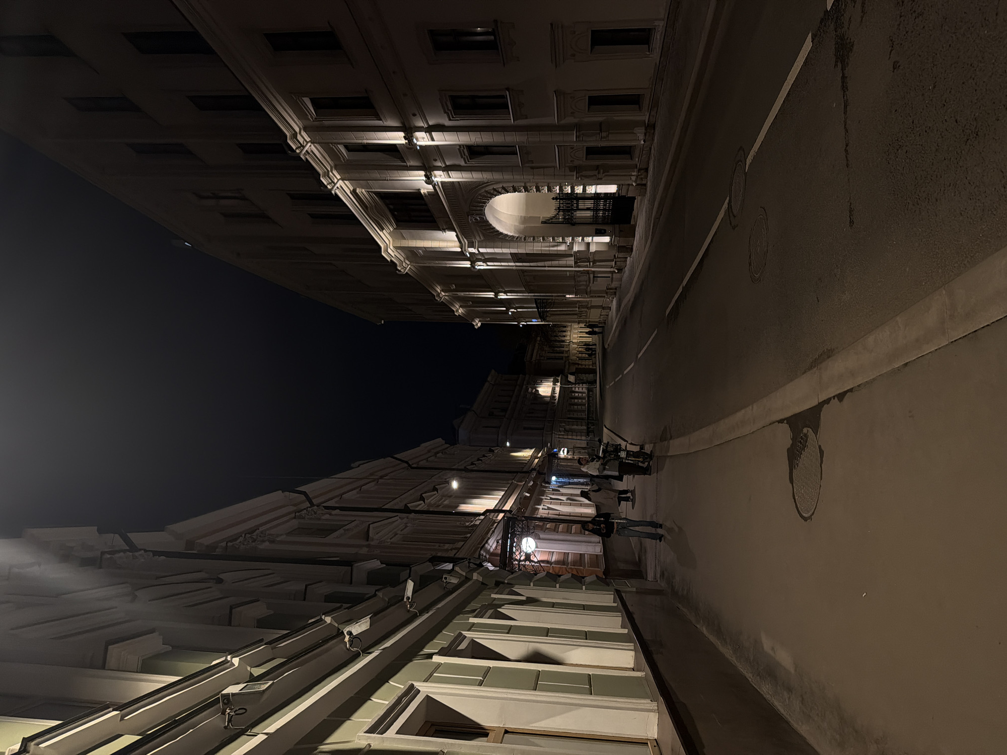 The image depicts a street scene in Moscow, Russia, at night. The buildings on either side of the street are illuminated, showcasing architectural details. People are walking along the sidewalk. The street is paved and includes features like manhole covers and painted lines. The sky is dark, and the lighting suggests it is after dusk.
