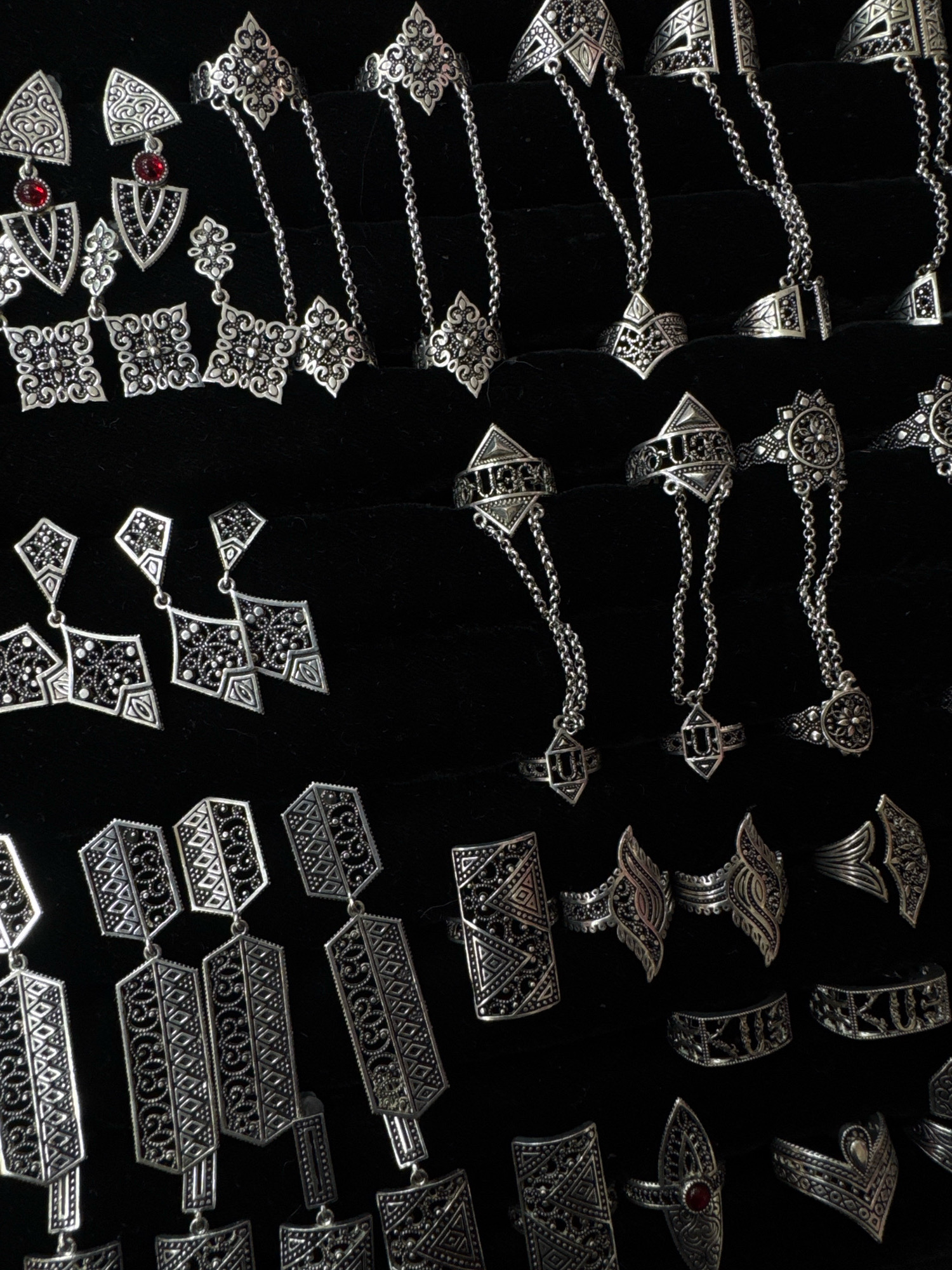 The image shows a close-up, top-down view of a collection of ornate, silver-toned jewelry displayed on a black fabric surface. The jewelry includes rings, earrings, and what appear to be hand chains or elaborate ring sets. The items are arranged in rows and columns, with many pieces exhibiting intricate filigree work, geometric patterns, and some with small red stones. The lighting is somewhat dim, creating a dark background that makes the metallic sheen of the jewelry stand out. There are no people visible in the image, and no specific location within the city of Ussurijsk, Russia, can be identified from the visual information provided. The scene is static and focuses solely on the display of the jewelry items.