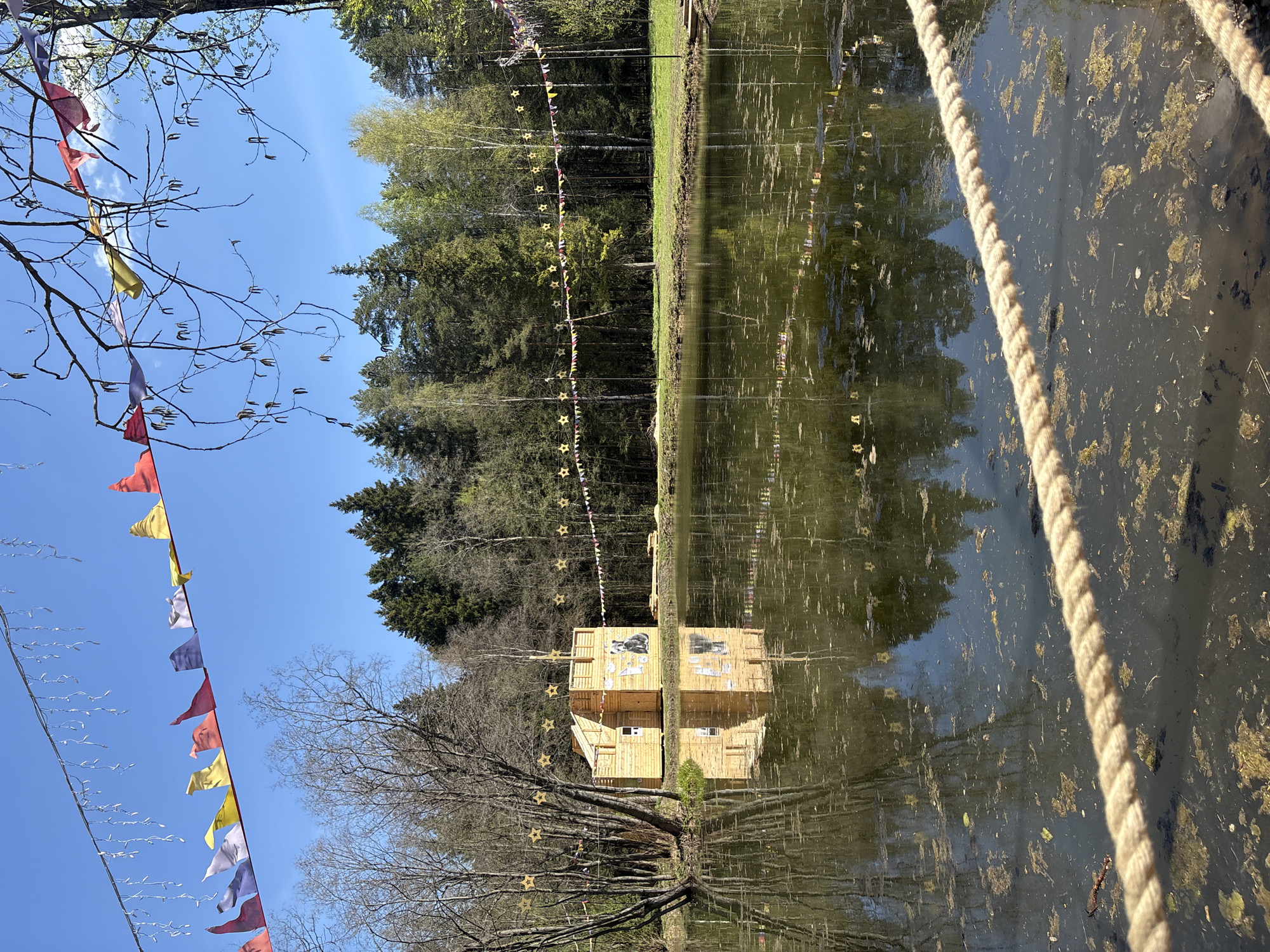 The image captures a landscape with a pond and a constructed wooden structure reflecting in the water. The scene includes trees, a blue sky with sparse clouds, and festive decorations such as colorful flags and strings of stars. A thick rope is visible on the right side of the frame. The wooden structure appears to be some kind of building or art installation. The location is Perm, Russia.