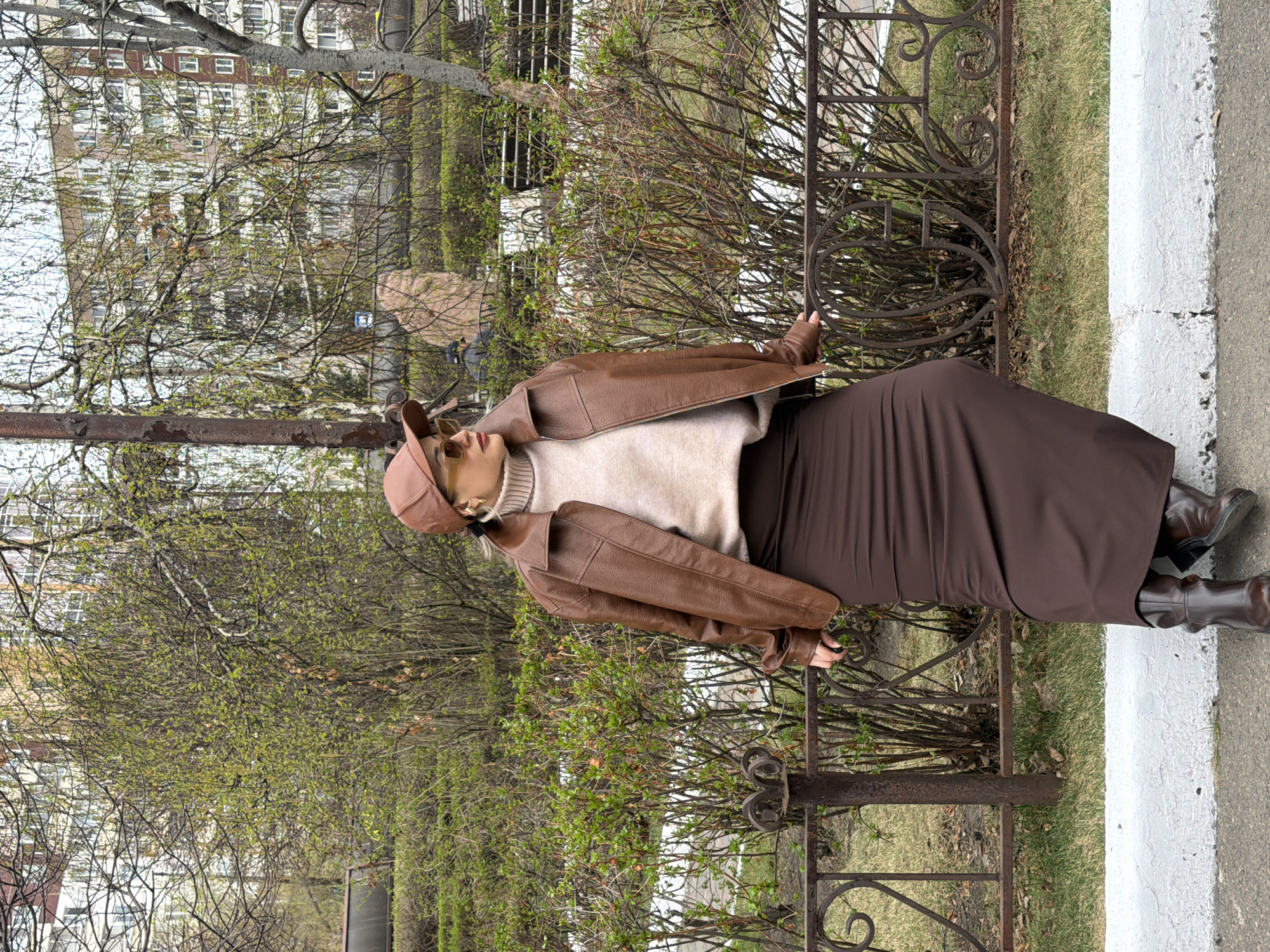 A woman stands beside a dark, ornate metal fence that borders a grassy area with shrubs in what appears to be an urban setting. She wears a beige turtleneck sweater, a brown leather jacket, a long brown skirt, and brown boots. A brown cap covers her head. Her hands rest on the fence. Behind her, foliage, possibly trees and bushes, are visible. Further in the background, a multi-story brick building is discernible. The scene is in Krasnoyarsk, Russia.