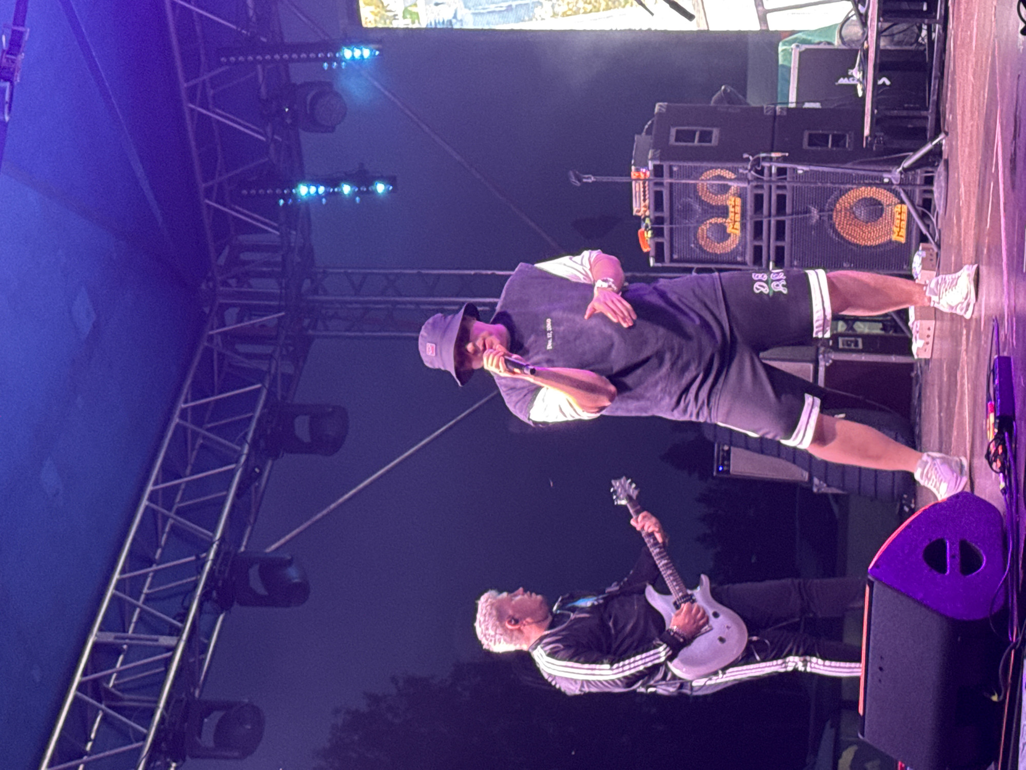 An outdoor music performance is depicted on a stage in Большая Кивара, Russia, under a dark, possibly nighttime sky illuminated by stage lighting. Two male performers are visible. The central figure, facing forward, wears a dark bucket hat, a dark sleeveless t-shirt with "Dec. 12, 2020" printed on the chest, dark shorts with white stripes and "DS AK" text on the left leg, and white sneakers. He holds a microphone to his mouth with his right hand and gestures with his left hand, wearing a watch. To his left and slightly behind him, a second male performer, with short light-colored hair, plays a light-grey electric guitar. He is dressed in a black tracksuit jacket with white stripes, black tracksuit pants with white stripes, and a light-colored necklace.
The stage features a metallic truss system above, from which several dark spotlights and blue string lights are suspended. On the right side of the stage, two large dark speaker cabinets are stacked, each featuring an orange circular speaker grille with a distinctive 'face-like' design. A smaller, purple-accented stage monitor speaker is on the floor in the lower left foreground. A microphone stand is visible behind the central performer, and various cables are strewn across the stage floor. Silhouetted trees are visible against the dark background on the left, and a large vertical screen displaying foliage is partially visible on the far right. The scene is predominantly lit with blue and purple stage lights.