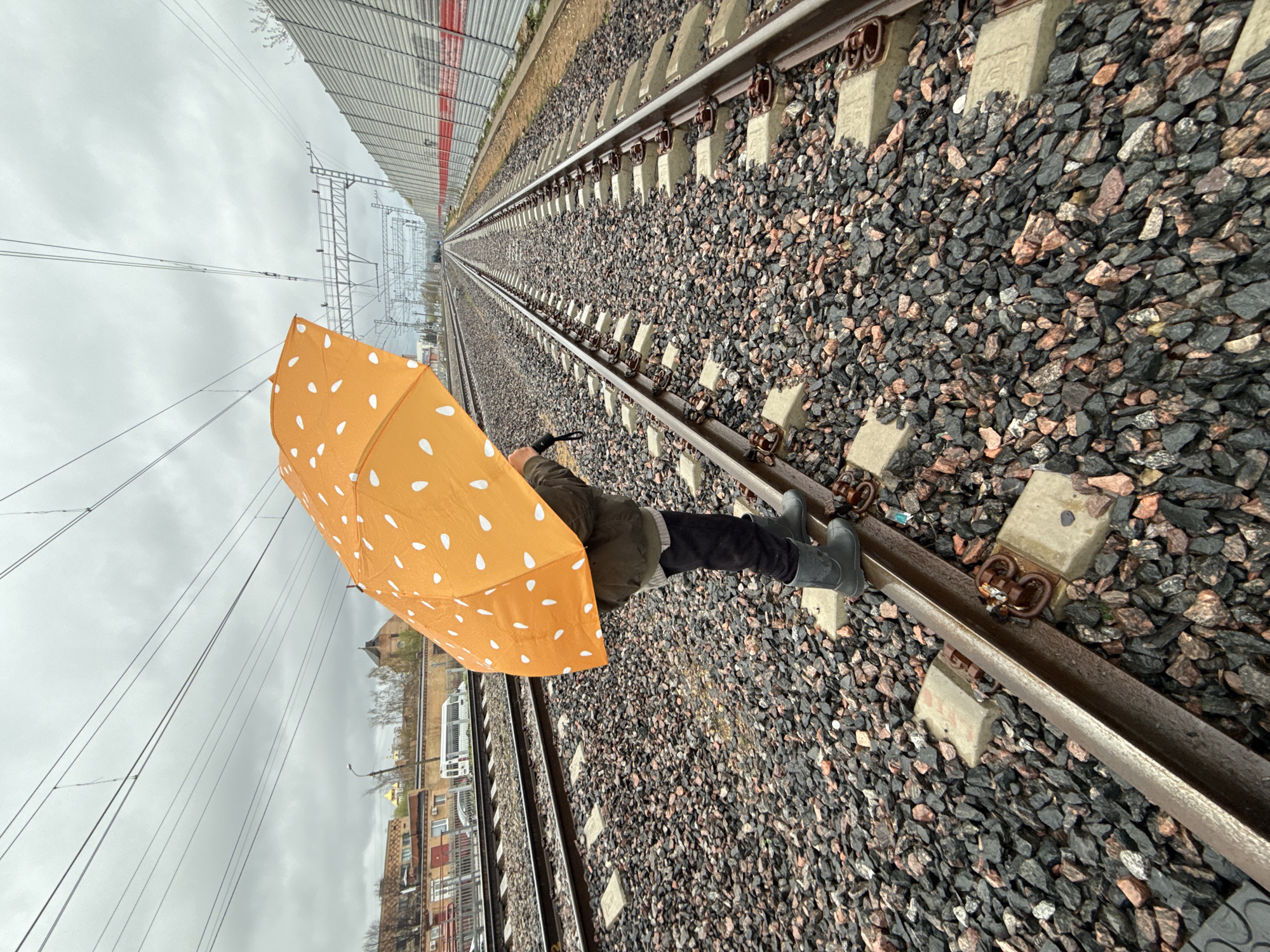 A person is standing on a railway track in Dmitrov, Russia. The person is holding a yellow umbrella with white polka dots and wearing a dark jacket and dark boots. The railway track extends into the distance, lined with gray stones. A tall fence with a red stripe runs parallel to the track on one side. The sky is overcast. Buildings are visible in the background.