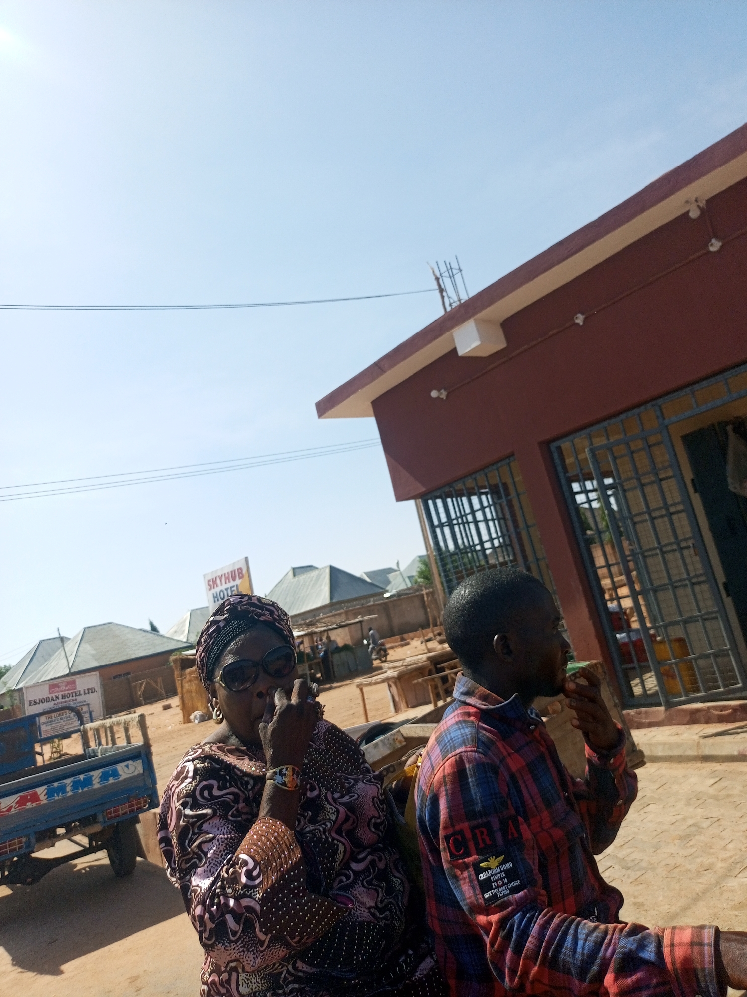 The image shows two people standing on a dirt road in what appears to be a small town. The woman on the left wears sunglasses, a patterned headwrap, and a patterned dress. She has her hand near her face. A man on the right is wearing a red and blue plaid shirt and also has his hand near his face.
Behind them is a building with a red exterior and barred windows. Further back, there are smaller buildings with corrugated metal roofs. A blue truck with white lettering is visible on the left side of the image.
A sign that reads "SKYHUB HOTEL" is seen in the background. Another sign is visible, bearing text that includes "ESJODAN HOTEL LTD. ADDRESS". The sky is clear and blue.