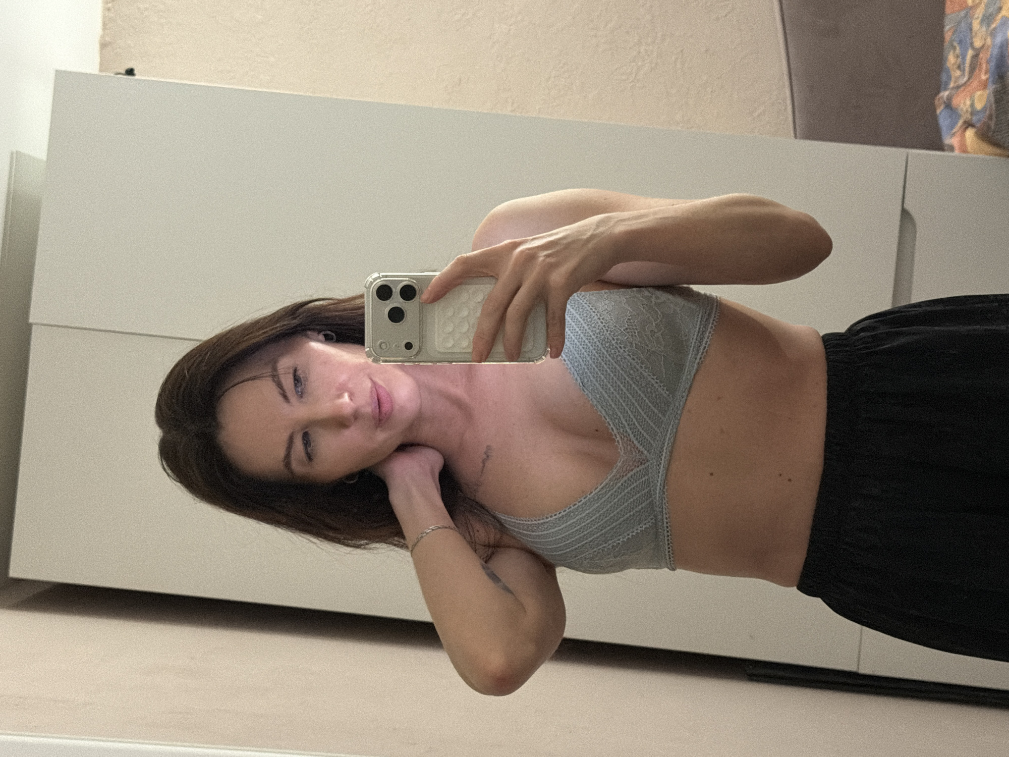 A woman is taking a selfie in what appears to be an indoor setting. She is holding a smartphone in her right hand, with her left hand resting near her neck. She is wearing a light blue bra and dark-colored bottoms. The background features a light-colored wall and a piece of furniture, possibly a dresser or cabinet. The image does not provide enough information to verify the location in the city of Perm, Russia.