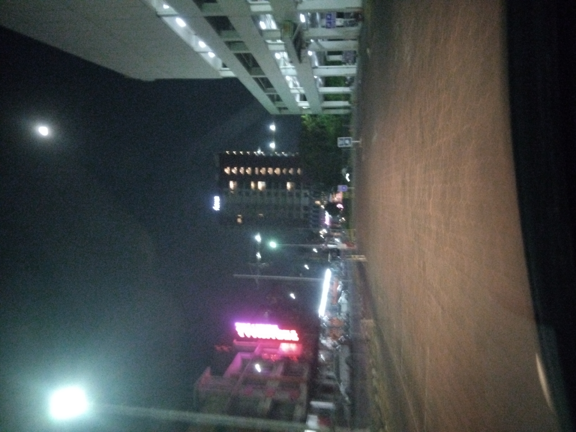 The image depicts a street scene at night in Surabaya, Indonesia. A bright street lamp illuminates the foreground, casting a glow on the asphalt road. To the left, a multi-story building with illuminated windows stands adjacent to a pink neon sign. Further down the street, a larger, darker building with more lit windows is visible. On the right side of the road, a multi-story concrete structure with open levels and visible lighting dominates the scene. The sky is dark with a faint, distant moon. The road appears to be paved with cobblestones or patterned concrete. The overall impression is one of an urban environment at night, with a mix of commercial and residential buildings.