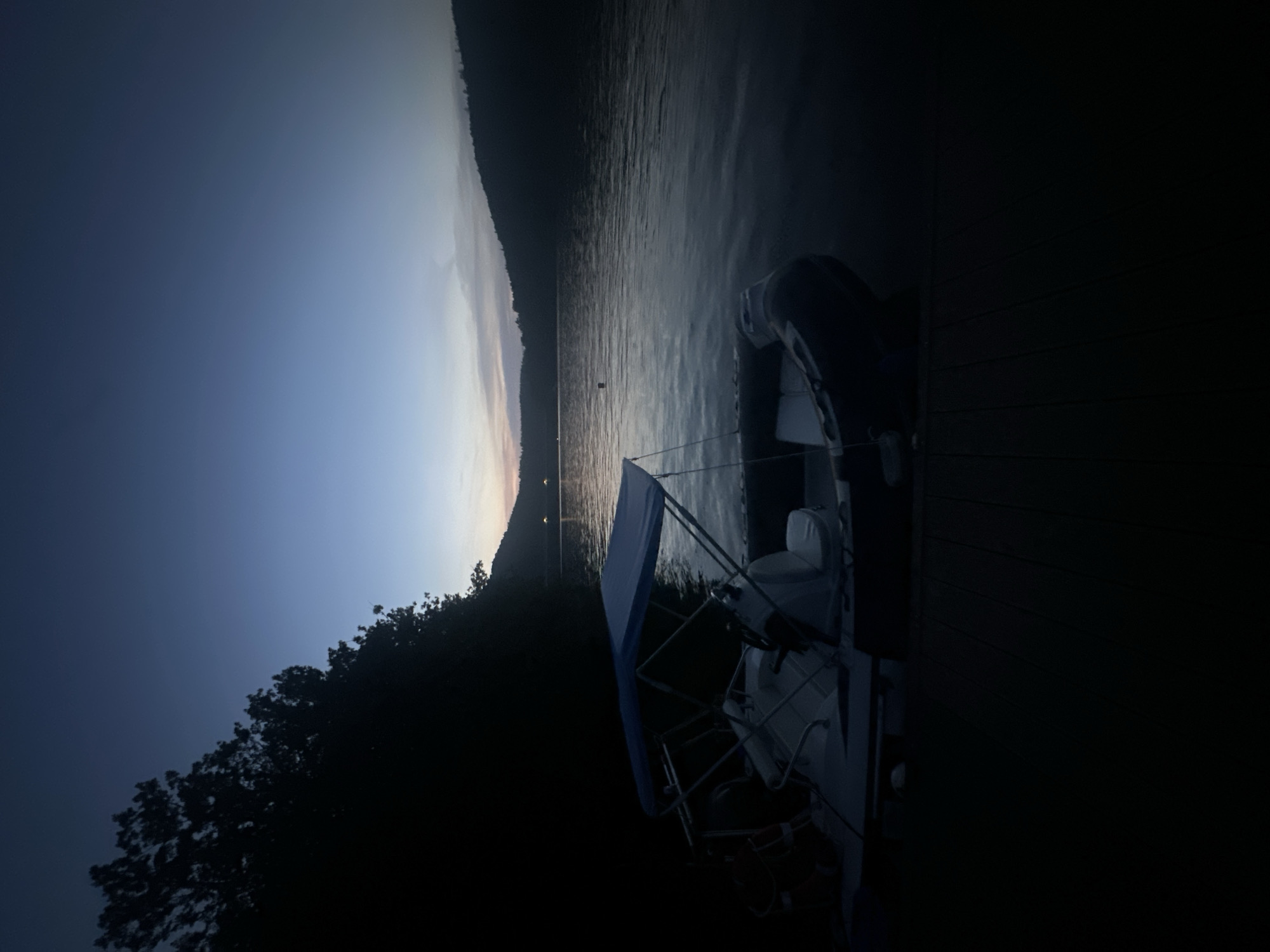 At dusk, a small, inflatable boat with a blue canopy is docked on a wooden pier. The boat is moored to the pier and the water is calm. The surrounding environment is dark, with trees on the left and a hill in the distance. The sky is a deep blue transitioning to lighter hues near the horizon, suggesting twilight. There are no people visible in the scene.
