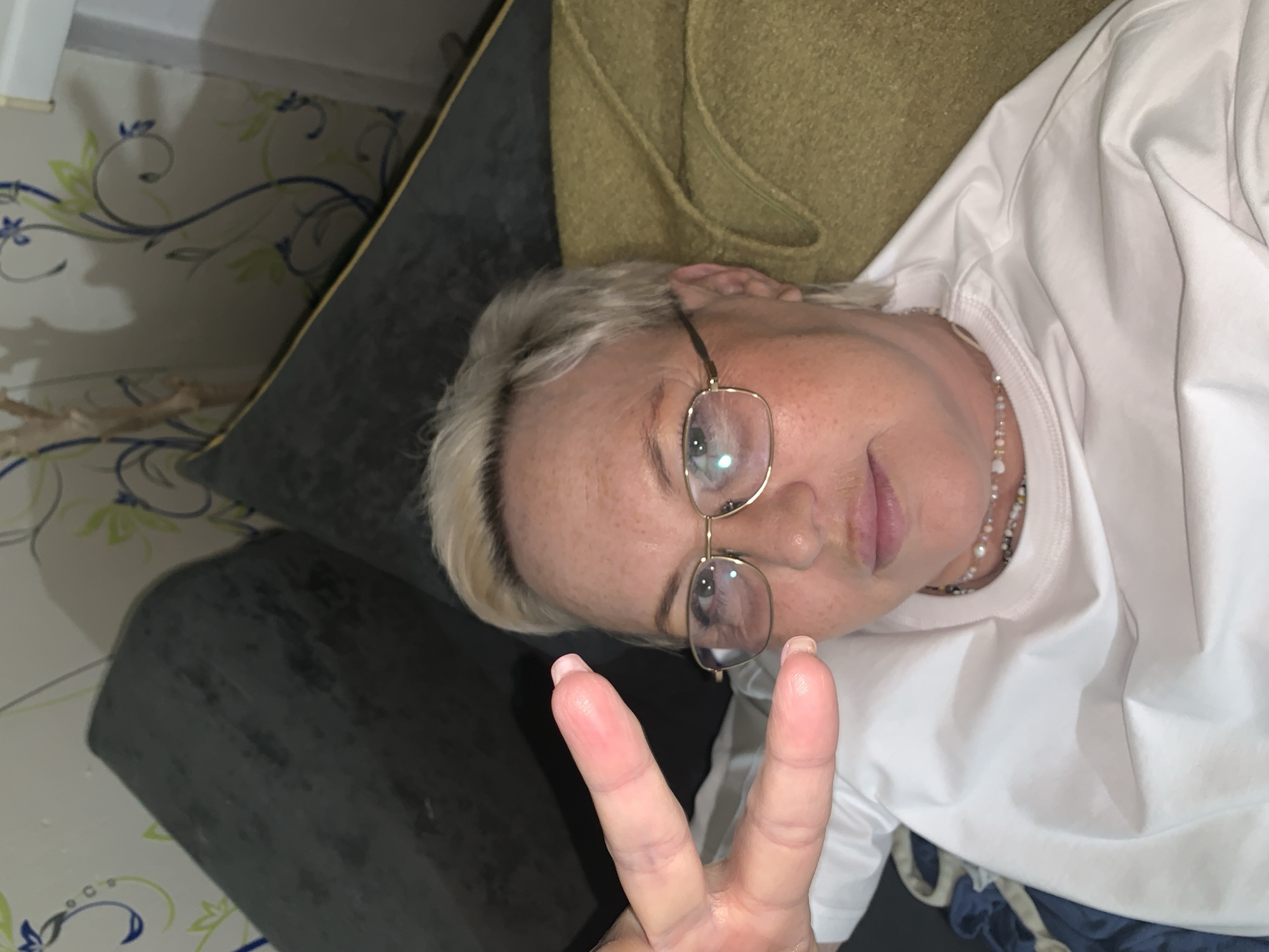 A middle-aged woman with short blonde hair, wearing gold-rimmed glasses and a white t-shirt, lies on a grey-green couch, making a peace sign with her hand. She is wearing a beaded necklace. The background includes a wall with a floral vine design. The location is an interior space, potentially in Chaikovsky, Russia.