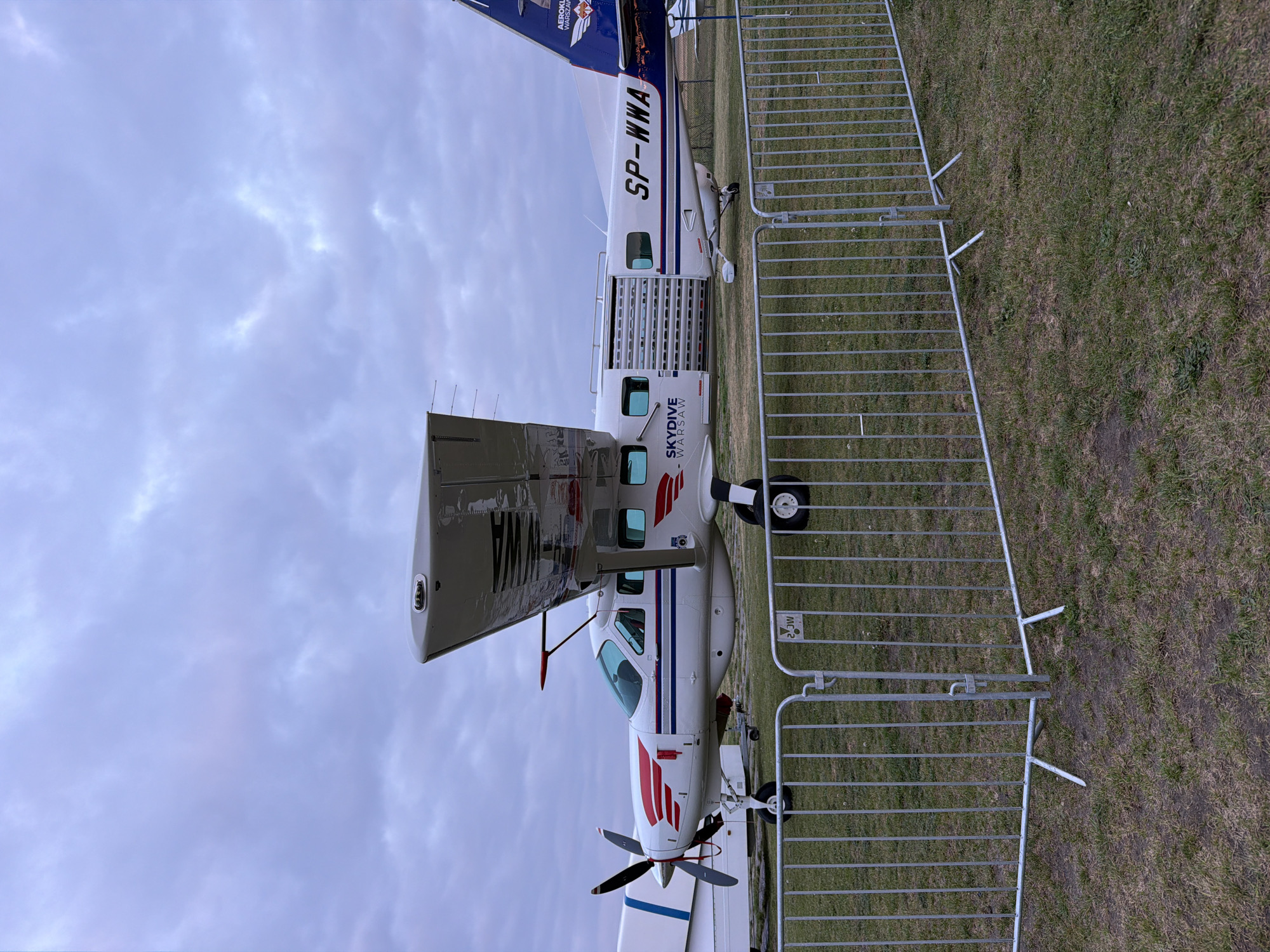 A sideways, low-angle shot captures a white and blue airplane on a grassy field. The aircraft has the words "SKYDIVE - WARSAW" written on its side. The plane's tail reads "SP-WWA." The propeller of the airplane is facing towards the bottom of the frame, and a silver metal fence is present on the right side of the image. The sky is a cloudy, pale blue.