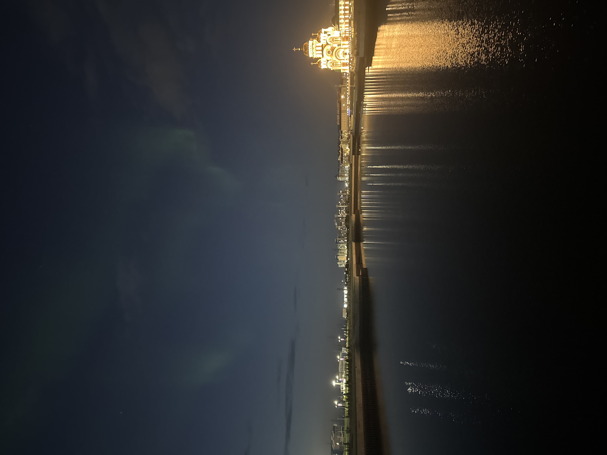 The image depicts a nighttime cityscape along a body of water, likely a river or bay, with the aurora borealis visible in the dark sky. In the distance, a large, illuminated building with golden domes and crosses, characteristic of a Russian Orthodox church, dominates the right side of the frame. The building's golden lights are reflected in the dark water. To the left of the church, a line of city lights indicates a developed urban area. Along the near bank of the water, there is a dark, textured retaining wall. The sky is a deep blue-black, with faint green streaks of the aurora and a few stars visible. The water's surface is mostly dark and still, with light reflections from the city and the church. The scene appears to be in Salekhard, Russia, a city known for its location above the Arctic Circle and its opportunities to witness the aurora borealis.