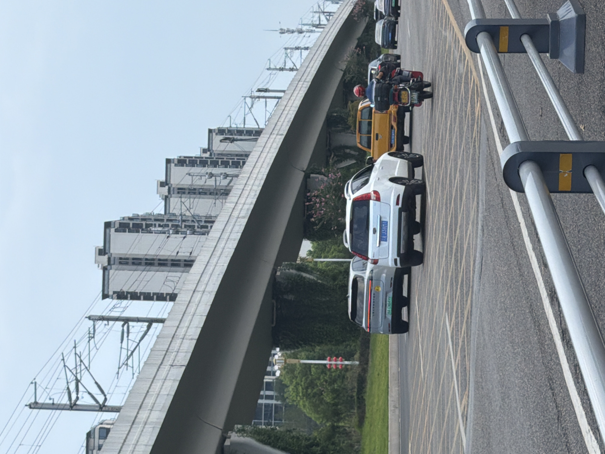 This image captures a scene on a multi-lane road under an elevated train or highway structure in Jiangjin, China. The sky is overcast. Several vehicles are present: a white SUV in the foreground with a license plate that appears to read "A J38M1", a silver SUV behind it with license plate "BA C11499", a yellow pickup truck, a black sedan, a motorcycle with a rider wearing a red helmet, and another black car further in the distance. The road surface is asphalt with yellow and white lane markings. The elevated structure is supported by concrete pillars, some of which are covered in greenery. Tall, modern apartment buildings with dark window frames are visible in the background, bordering the elevated structure. Overhead power lines for an electric train are strung between poles along the elevated structure. The overall environment suggests a busy urban setting.