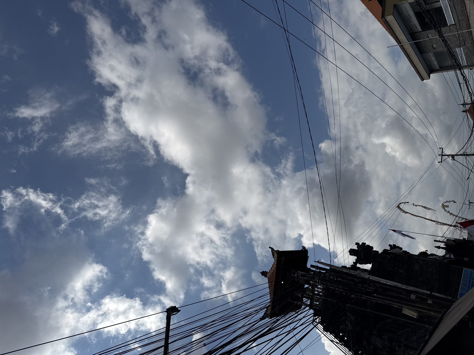 The image shows a view looking upwards towards a cloudy blue sky, with a traditional Balinese structure and modern buildings visible. A dark, ornate Balinese structure, possibly a temple gate or monument, is silhouetted on the left side of the frame. It has a tiered roof with intricate carvings and is surrounded by a dense network of black electrical wires. A small Indonesian flag is visible on a pole next to this structure, along with decorative festive offerings. To the right, a section of a modern building with a tiled orange roof and grey stone facade is visible, also entangled with power lines. The overall scene suggests an urban environment in Denpasar, Indonesia, where traditional architecture and religious elements coexist with modern infrastructure and a bustling electrical grid. The lighting indicates it is daytime, with sunlight illuminating the clouds and creating some shadows.
