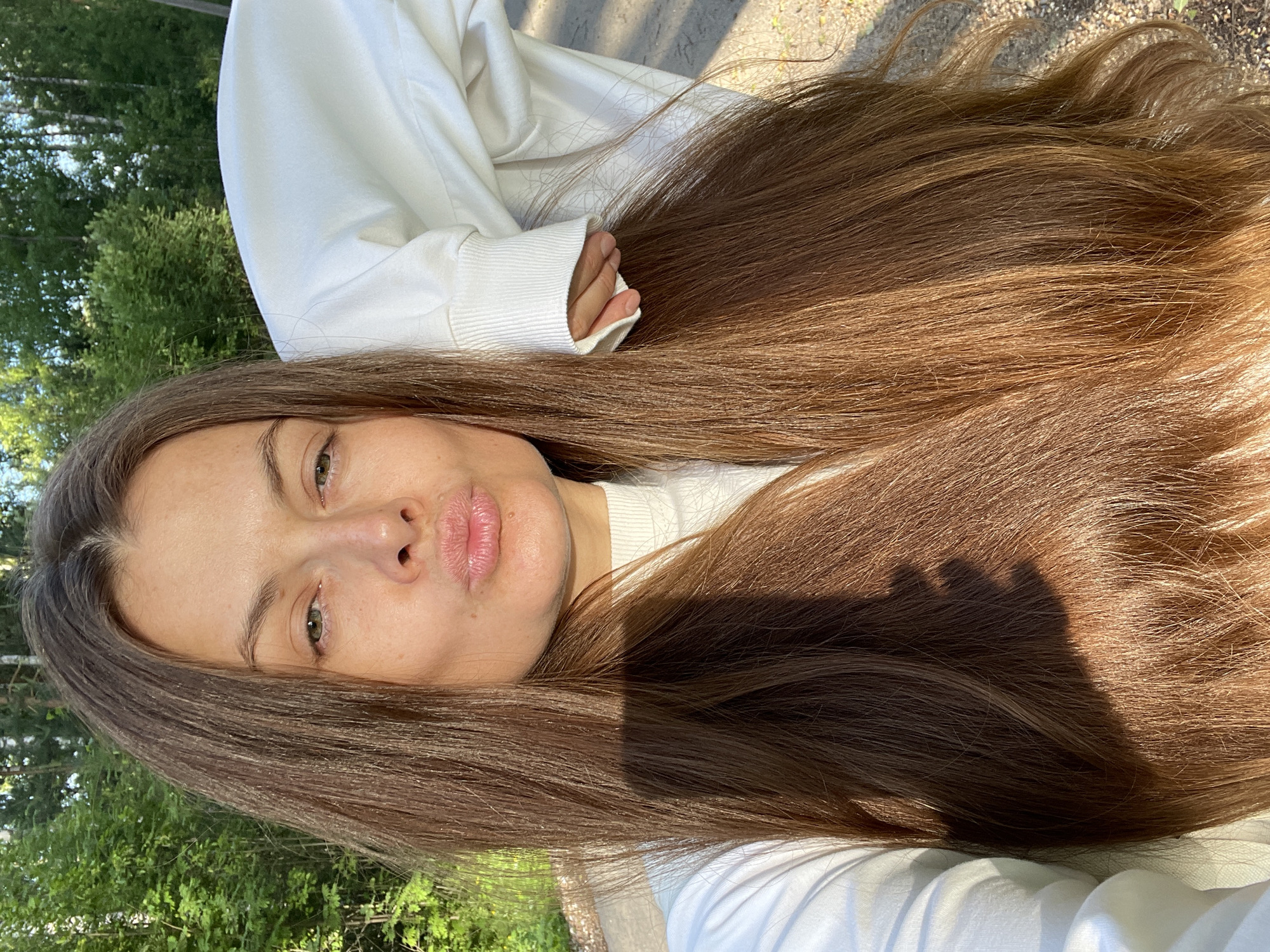 A woman with long brown hair is taking a selfie outdoors. She is wearing a white turtleneck and a white sweatshirt. Her lips are pursed, and she has a shadow cast on her hair. The background consists of trees and a gravel path, suggesting a park or wooded area. There are no identifiable landmarks to pinpoint the specific location within Заречный, Russia.
