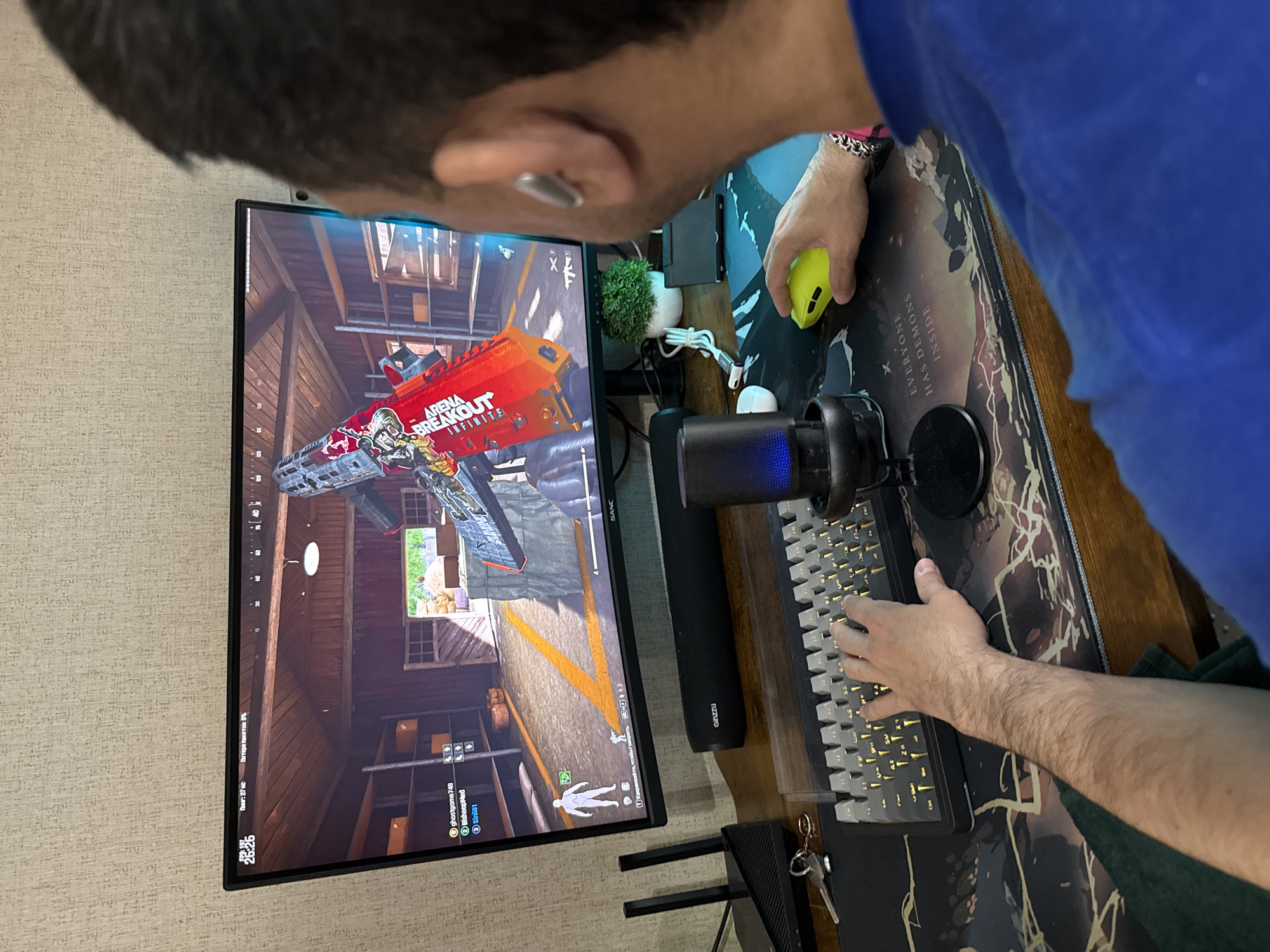 A person is playing the game "Arena Breakout" on a computer monitor. The screen displays a first-person view inside a wooden structure with a gun model prominently visible. The player is using a yellow mouse and a mechanical keyboard with yellow backlighting. A microphone, speaker, and a small potted plant are also on the desk. The environment consists of a textured wallpaper, a decorative mousepad, and an Xbox console. The location is likely indoors and not possible to be determined in the city of Rusanovo, Russia, based solely on this image.