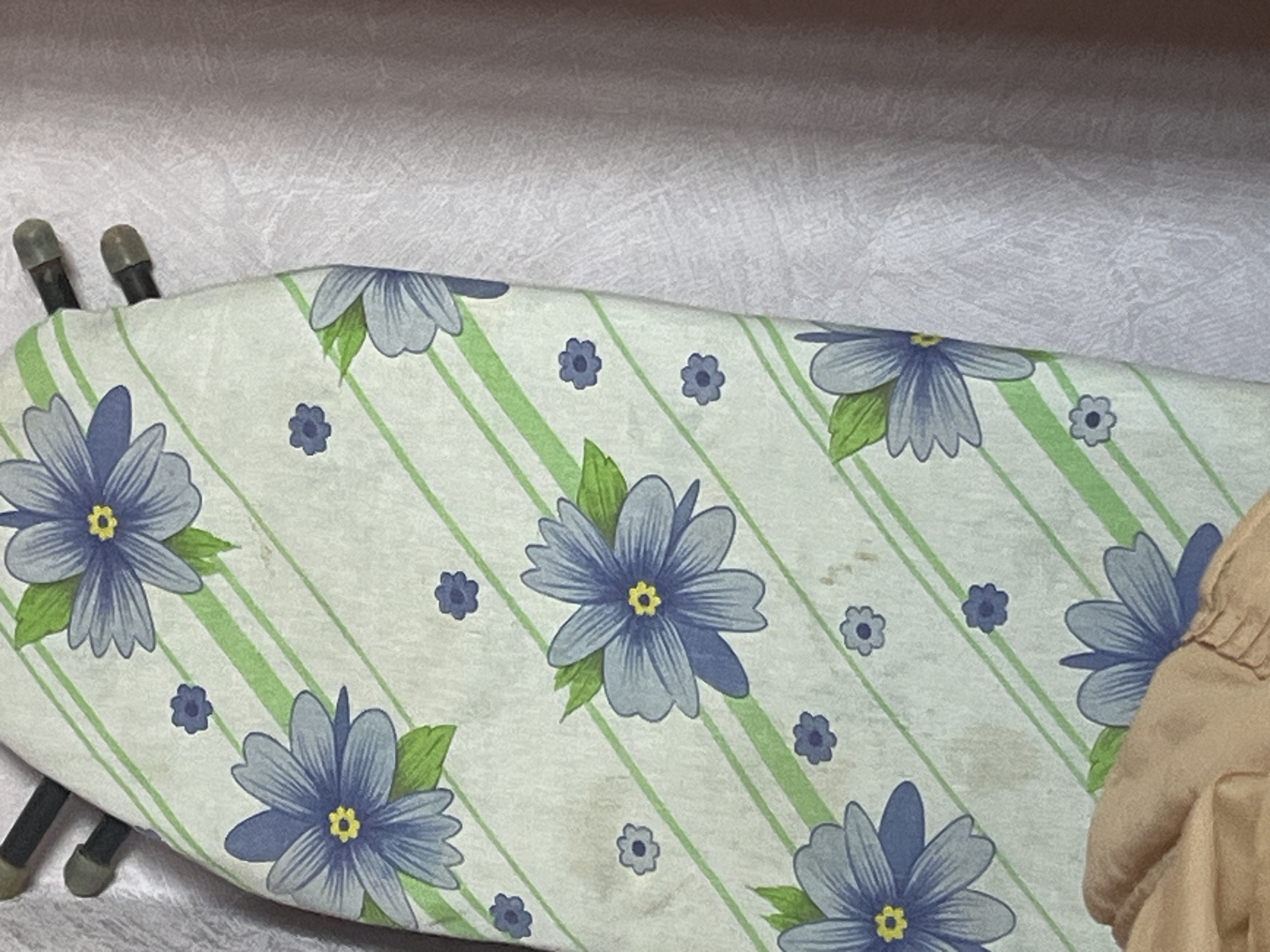 The image shows a close-up of an ironing board cover with a pattern of blue flowers and green stripes. The ironing board is leaning against a light purple textured fabric, possibly a curtain or wall covering. Two dark rods with grey caps are visible at the top edge of the ironing board. A portion of a person's arm or shoulder, covered in beige fabric, is in the lower left corner of the frame. There are also some brown stains on the ironing board cover. The location in the city of Gari, Russia is not discernible from the visual information provided in the image.