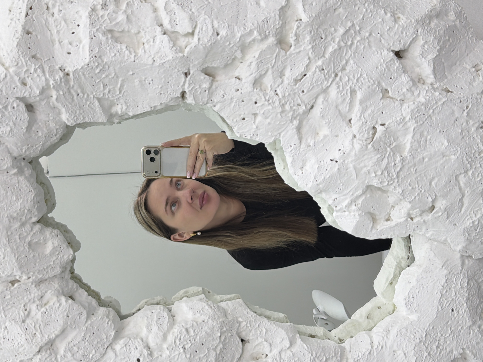A woman is reflected in a mirror, holding a smartphone in her right hand, presumably taking a selfie. She has light brown hair with blonde highlights and is wearing a black top. The mirror is irregularly shaped, with a thick, textured white frame resembling stucco or plaster. The background, as seen in the mirror, appears to be a plain white wall. The location is identified as Moscow, Russia, though this information is not explicitly depicted in the image.