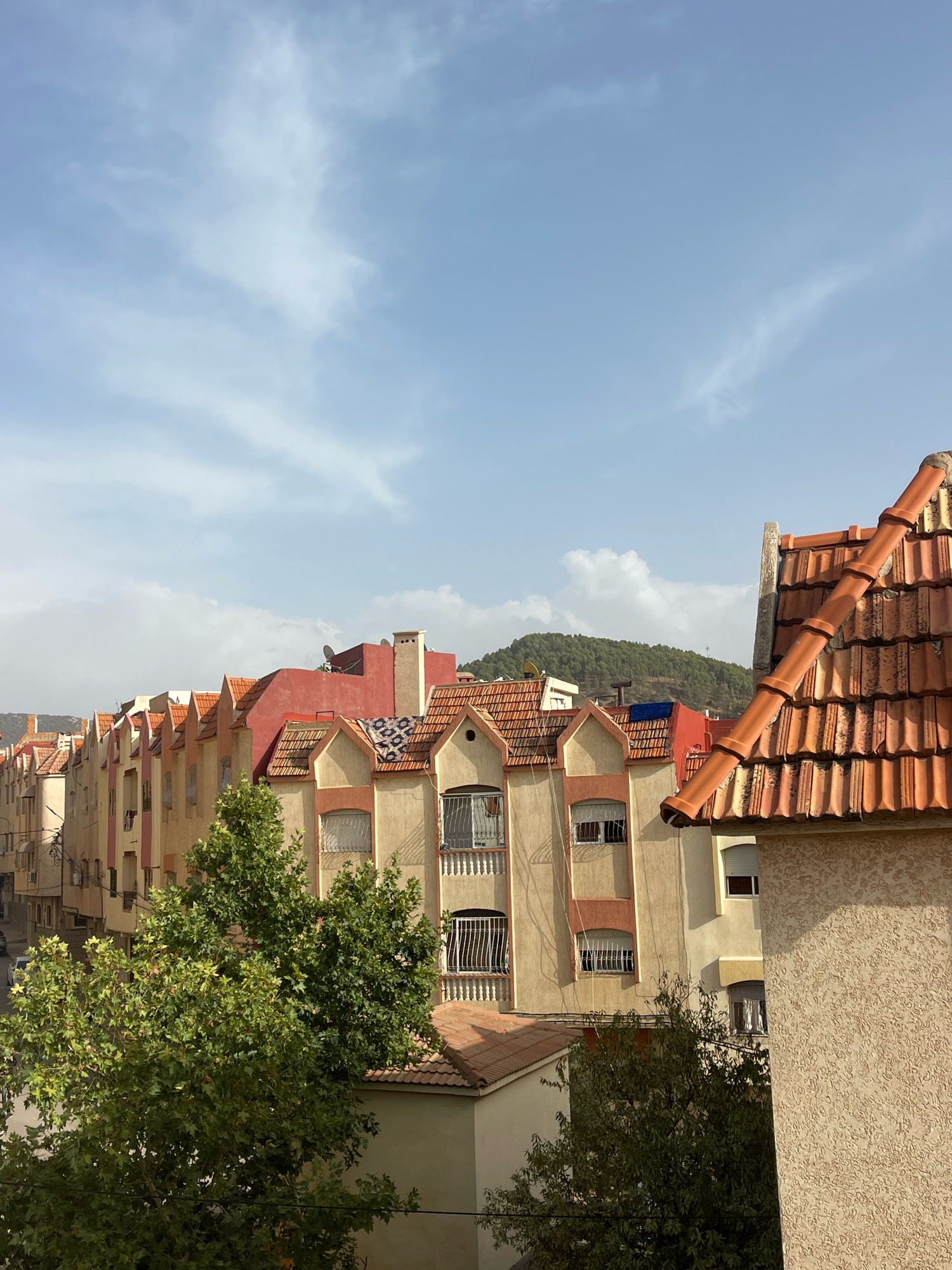 A medium shot captures a street scene in Azrou, Morocco, under a partly cloudy sky. The foreground features a prominent, textured beige wall on the right, with an orange tiled roof extending from the top right corner diagonally across the frame. To the left of the beige wall and below the roof is another lower orange tiled roof.
Beyond these elements, a row of attached buildings with reddish-orange tiled roofs lines the street. These buildings display a mix of architectural styles and colors, predominantly in shades of yellow, beige, and red. Many of the buildings have decorative gables and some feature balconies with white railings. Trees with lush green foliage are interspersed between the buildings, partially obscuring some of the lower floors. In the background, a green, tree-covered hill rises under the sky. The overall impression is of a residential area with a distinct Mediterranean or North African architectural character.
