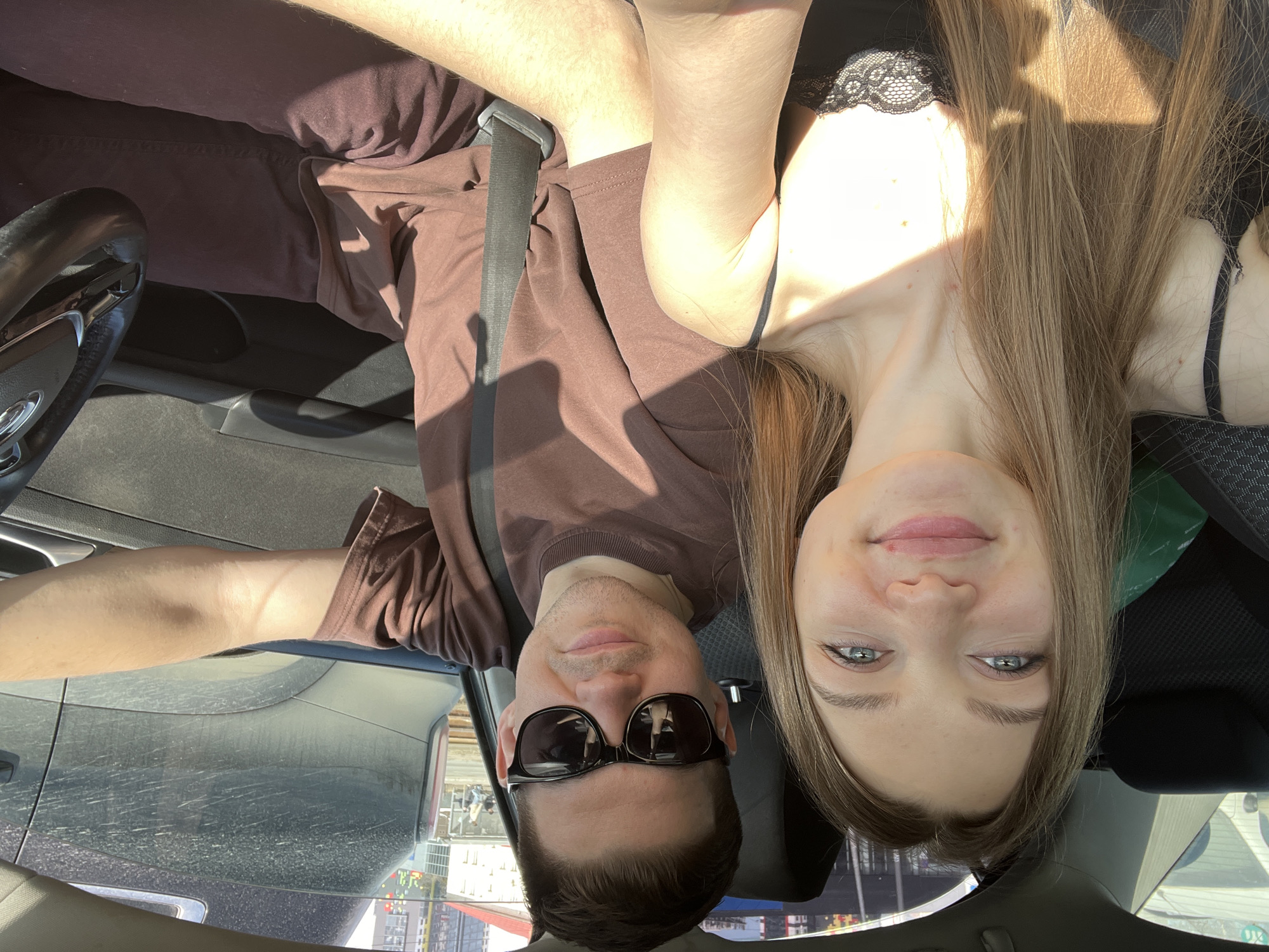 The image depicts two individuals inside a car. A female, with long light brown hair and blue eyes, wearing a black lace-detailed top, occupies the front passenger seat, holding a device to capture a selfie-style photograph. A male, wearing a brown t-shirt, dark trousers, and dark sunglasses, occupies the driver's seat, with his left hand resting on the steering wheel and a dark seatbelt fastened across his chest. Both individuals present faint smiles, looking towards the camera. Strong sunlight enters the vehicle, casting defined shadows on the occupants and interior.
Through the driver's side window, an urban streetscape is visible, featuring a dusty dark-colored vehicle in an adjacent lane and tram tracks running parallel to the road. In the background, modern multi-story buildings are present, including one with a distinctive facade characterized by green, blue, and red pixelated patterns. This architectural detail and the urban infrastructure place the scene in Kazan, Russia, specifically near the **IT Park** or the **Universiade Village** area, adjacent to a thoroughfare with tram lines.