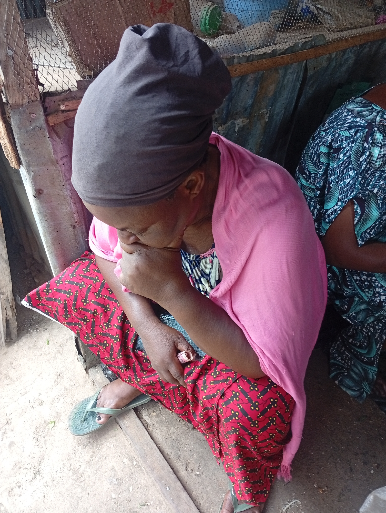 An adult female sits in a ground-level exterior space in Sabon Tasha, Nigeria. She wears a dark gray head wrap, pink shawl, and red and black patterned pants. She has green flip-flops on her feet. She is seated near a person with a teal and navy patterned top. Her face is partially covered by her hand. The ground is dirt. A wire cage is visible behind her.