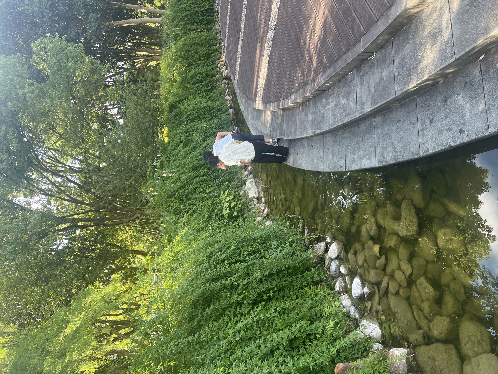 The image depicts a tranquil scene in Liandu District, China, featuring two young individuals, a boy and a girl, by a shallow stream. The boy, wearing a white shirt with a floral pattern and black pants, is leaning over the water's edge, as is the girl, who is wearing a white and blue striped shirt and dark shorts. They appear to be observing something in the stream.
The stream itself is clear, revealing a bed of smooth, grey and white stones. Lush, green foliage drapes over the stream bank, creating a natural, verdant border. To the right of the stream, a paved walkway with a distinctive wooden plank pattern curves away, suggesting a well-maintained public space. The overall environment is serene and natural, with sunlight filtering through the trees, casting dappled shadows on the walkway.