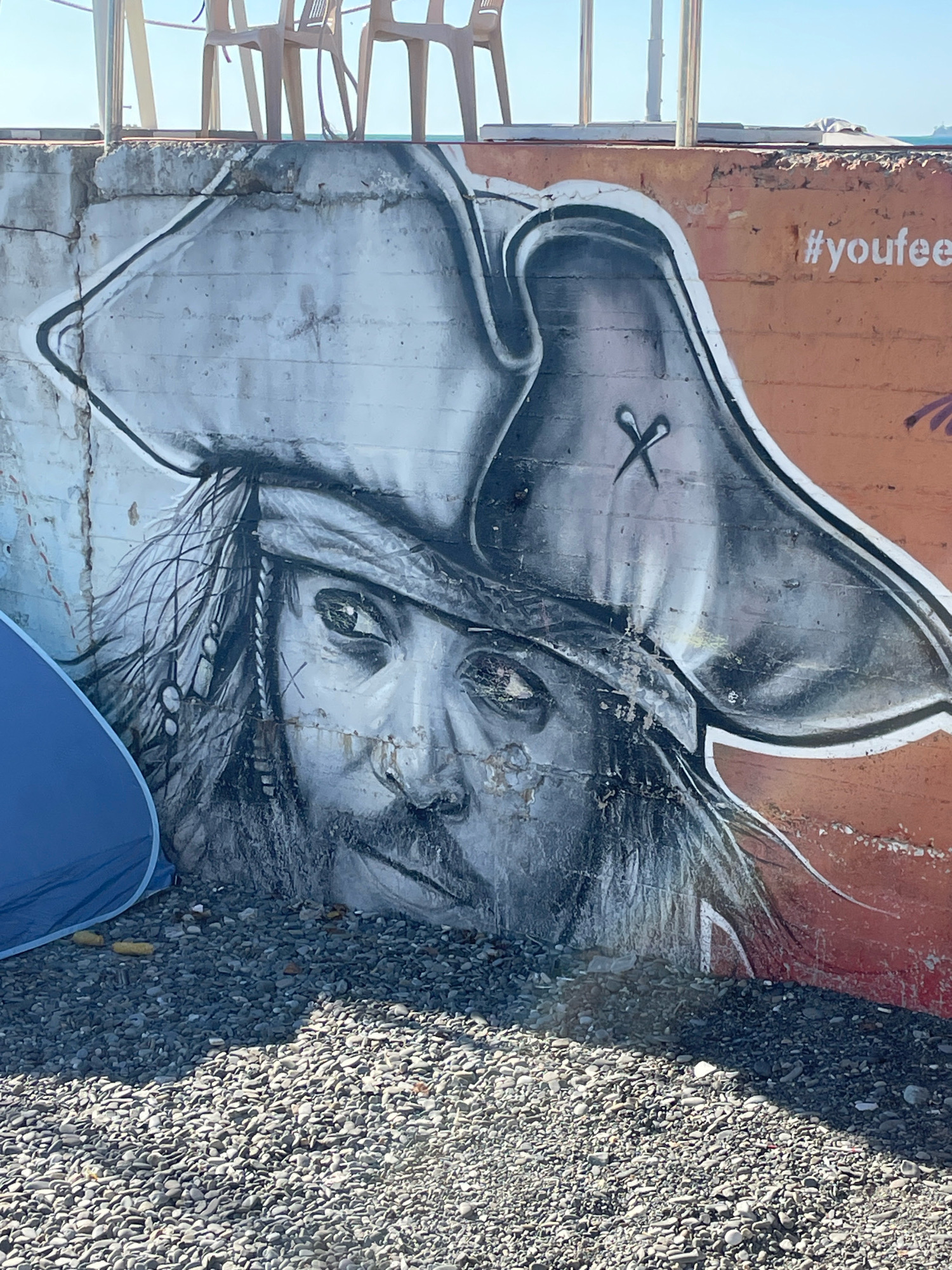 This image depicts graffiti on a concrete wall near a beach in Sochi, Russia. The graffiti is a black and white portrait of a pirate with a large tricorn hat, reminiscent of Jack Sparrow. The pirate has detailed facial features, including an eye patch and beard. The word "#youfee" is spray-painted in white on the orange section of the wall to the right of the portrait. In the foreground, there is a patch of grey pebbles, typical of a beach. A blue tent is partially visible on the left side of the image, and in the background, several empty chairs are stacked near a railing, with the sea and sky visible beyond. The concrete wall appears weathered and cracked in places.