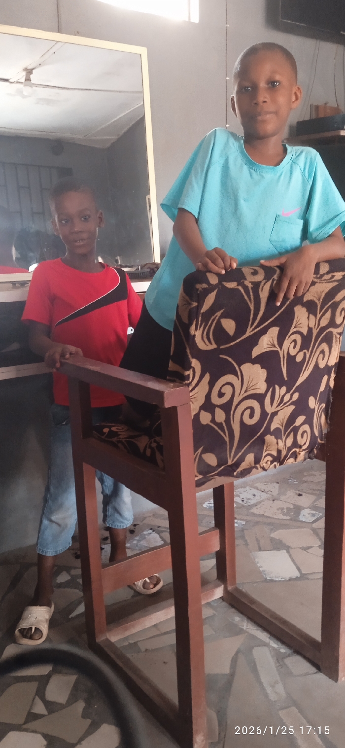 Two young boys pose beside and behind a wooden chair with a brown floral cushion. The boy on the left wears a red shirt with black accents and jeans, while the boy on the right sports a light blue t-shirt. Both are wearing sandals. A mirror is visible behind them. The room appears to be indoors, possibly a shop, with a tiled floor. A date stamp, "2026/1/25 17:15," is visible in the bottom right corner of the image.