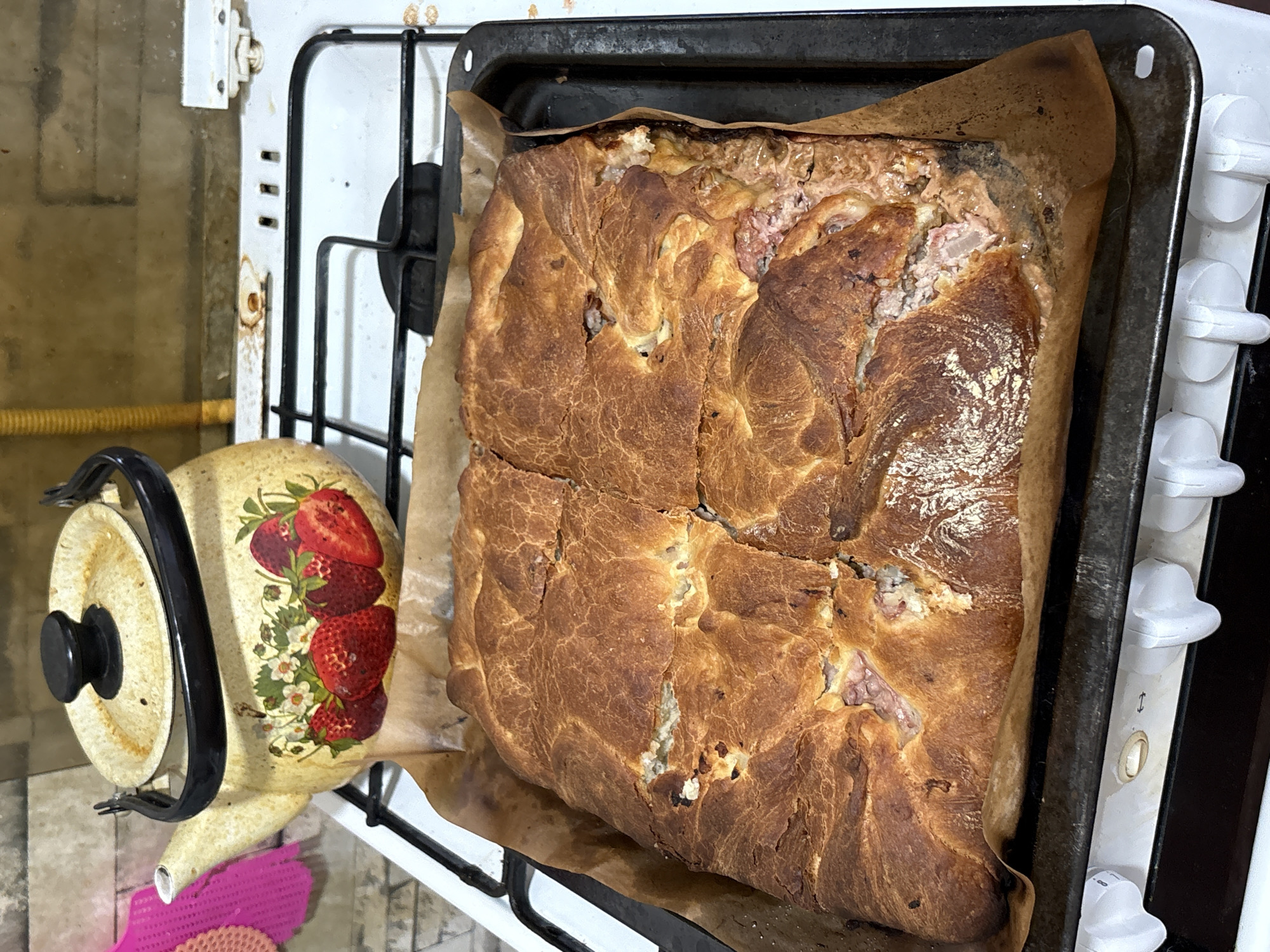 A close-up, eye-level shot shows a square, golden-brown baked pie on a dark baking sheet lined with parchment paper. The pie's crust is cracked and uneven, revealing pieces of its filling, which appears to be a mix of meat and possibly some sort of sauce or cheese. The baking sheet rests on a white gas stove. To the left of the pie, a cream-colored metal teapot with a black lid and handle is positioned on the stove. The teapot is decorated with a pattern of strawberries and white flowers. In the background, a brick wall is visible, with some lighter-colored tiles or grout between the bricks. The overall environment suggests a kitchen setting. There are no people visible in the image. The scene is not specifically identifiable as being in Shymkent, Kazakhstan, or any other specific city.