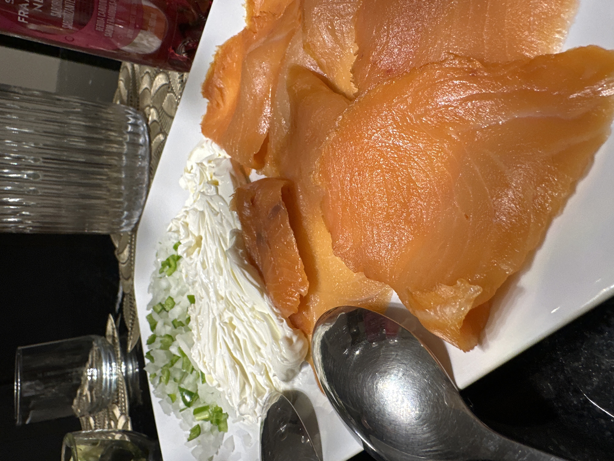 A close-up shot displays a white plate containing slices of smoked salmon, a creamy white spread sprinkled with diced green onions, and two spoons, set on a black countertop. The composition is framed from an elevated angle. A glass object and a bottle with red liquid are visible to the left of the plate. The photo appears to be taken in an indoor setting with artificial lighting. While the culinary nature of the presentation hints at a dining setting, there's no contextual information from which to infer a location in Mexico City.