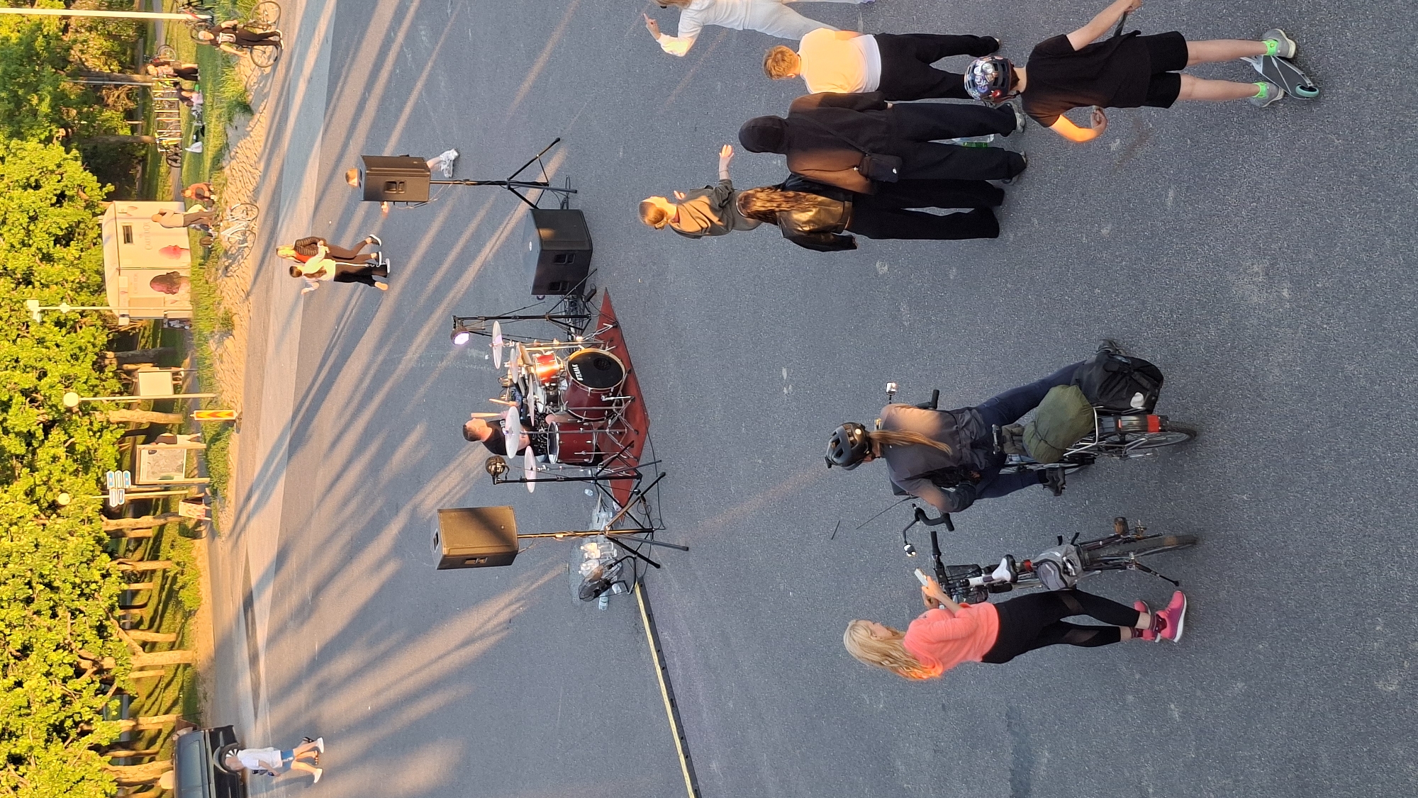 This aerial photograph, taken in Tallinn, Estonia, depicts an outdoor scene during daylight, likely late afternoon or early evening, given the long shadows cast by trees and structures. The foreground features two individuals on bicycles. A woman in a coral-colored top and black leggings stands next to a bicycle. To her right, another person, wearing a dark jacket and blue jeans and a helmet, is seated on a bicycle with luggage attached to the rear rack. Further into the midground, a drummer is actively playing a drum set, with two large speakers positioned nearby. Several other individuals are gathered around the drummer, some appearing to be watching the performance. In the background, there is a park-like area with trees and a building that appears to be a kiosk or small shop. Other people are visible in this area, some sitting on grass and others walking. The overall setting suggests a public gathering or event in a park or open space near the coast or a waterfront area.