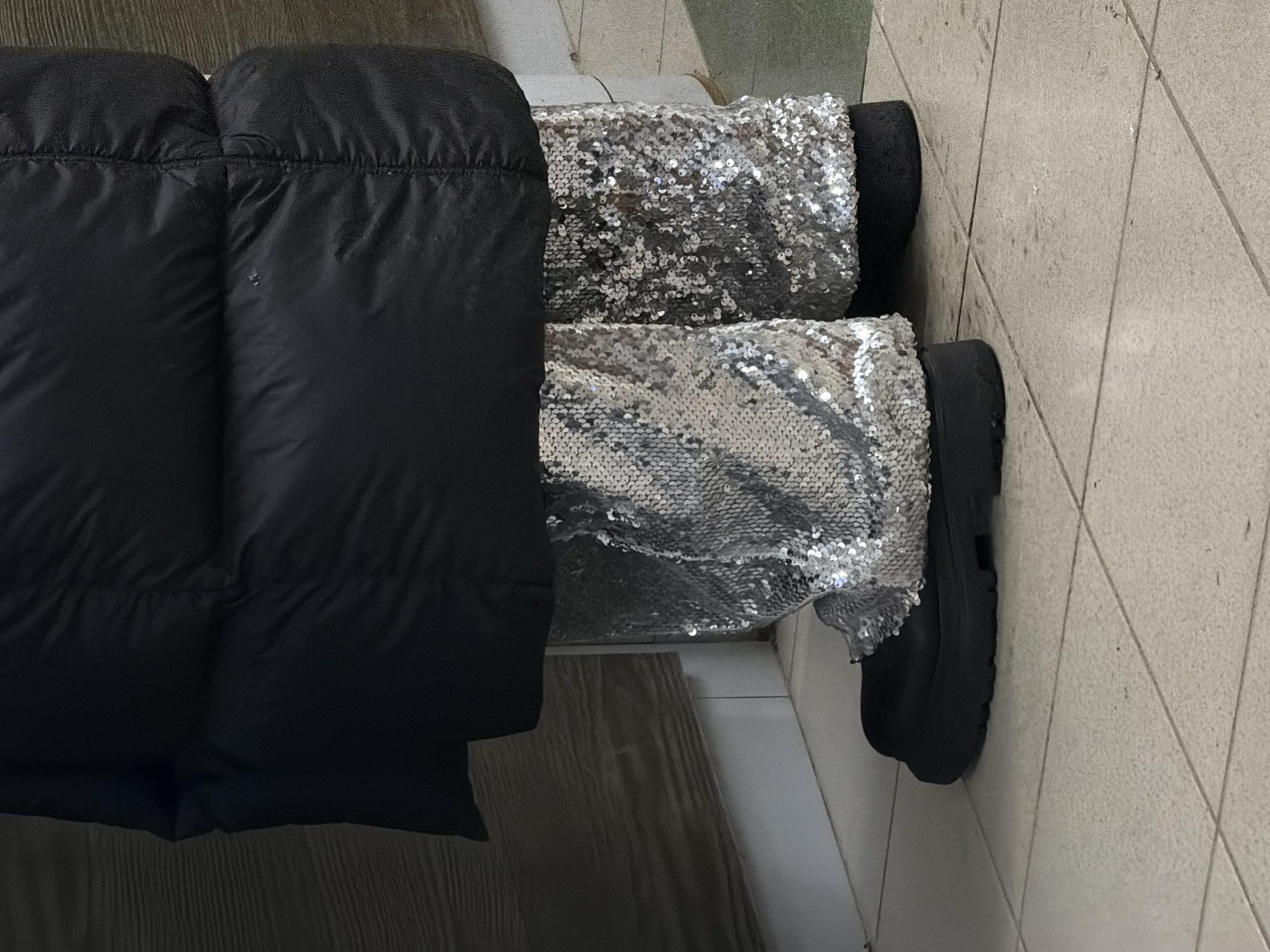 A puffy black coat covers the upper body of a person wearing silver sequined pants. The person wears black boots and stands on a tiled floor. The location is unknown.