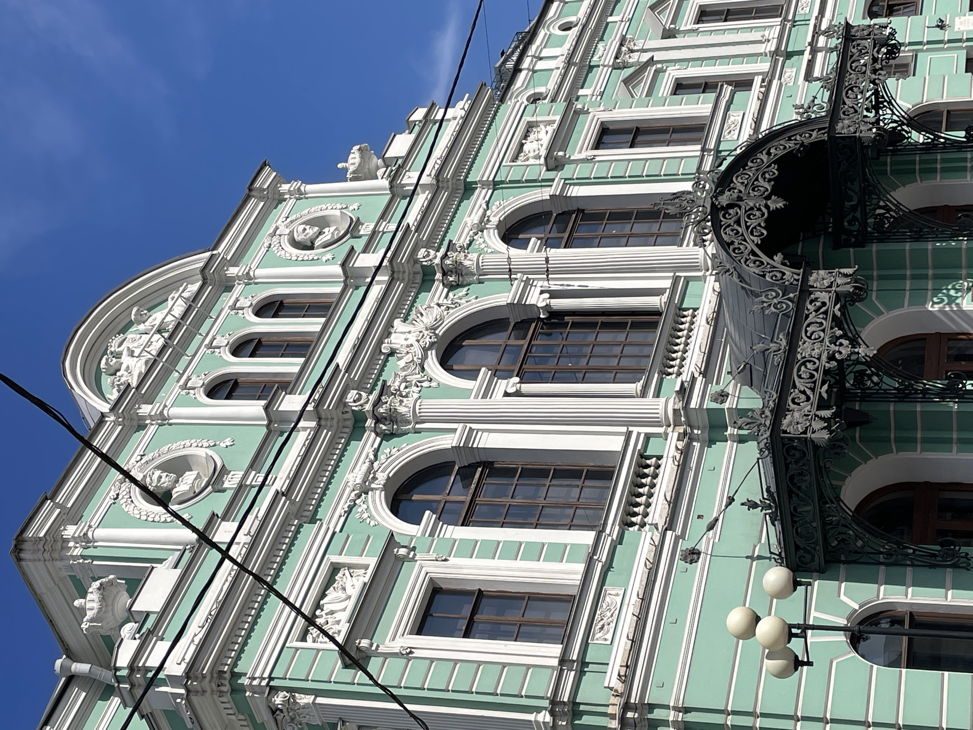 The image depicts a section of a building facade in Saint Petersburg, Russia. The building features a pale green and white color scheme with ornate architectural details. Elements include arched windows, sculpted figures, decorative columns, and an elaborately wrought iron balcony structure. A black electrical wire runs diagonally across the foreground. The sky is blue with scattered clouds.