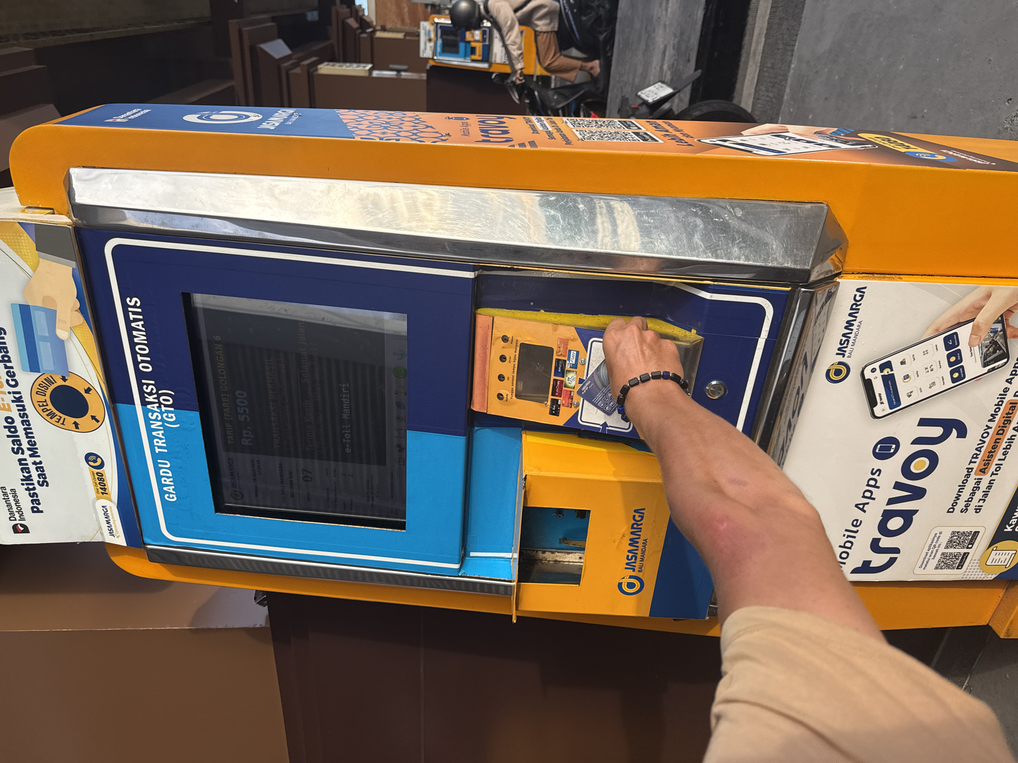 The image shows a person using an automated toll booth machine in Kuta, Bali, Indonesia. The machine is yellow and blue with the logo of "JASAMARGA BALI MANDARA." The screen displays information about the toll transaction, including the toll tariff which is 5500 Rp. The person's arm and hand are visible as they insert their e-toll card into the machine. The toll booth is in a garage. A motorcycle is parked in the background.